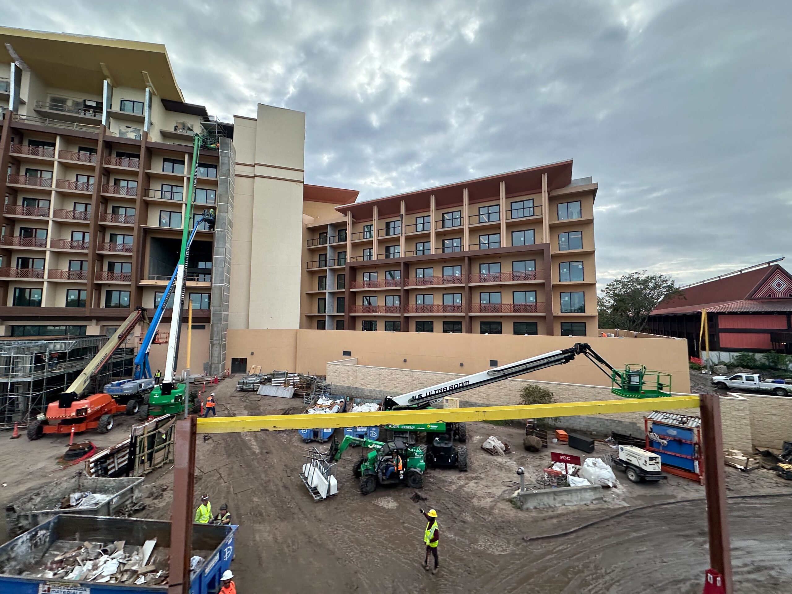 Construction site with workers and machinery in front of a multi-story hotel building. The sky is cloudy, and various construction materials are scattered around the ground.