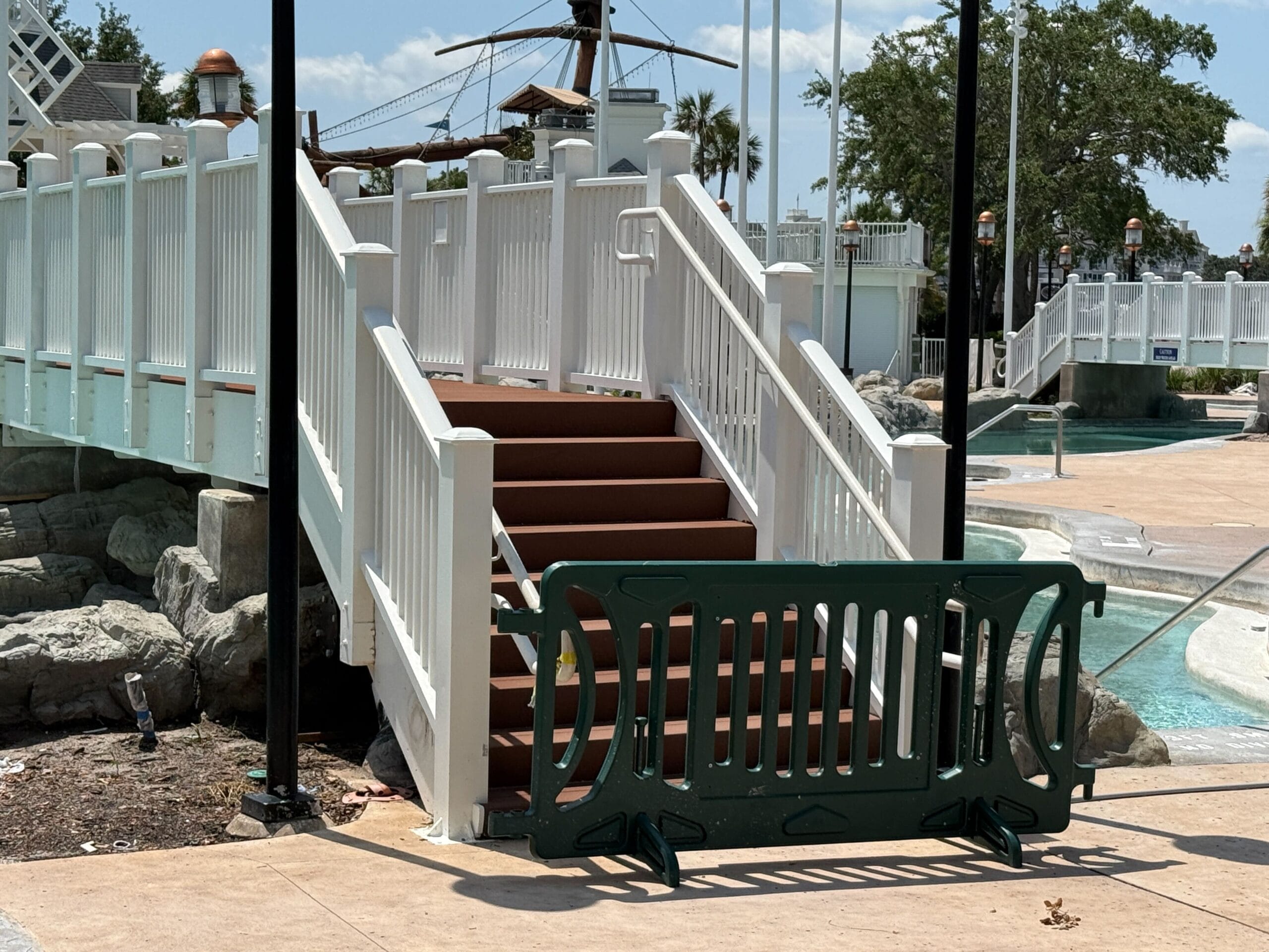A white stairway is blocked by a green barricade near Stormalong Bay’s ship-themed playground under a sunny sky.