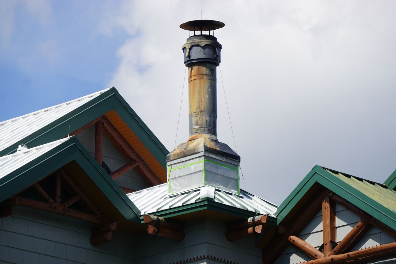 A tall, weathered chimney rises from a metal roof with green trim and wooden beams under a partly cloudy sky.