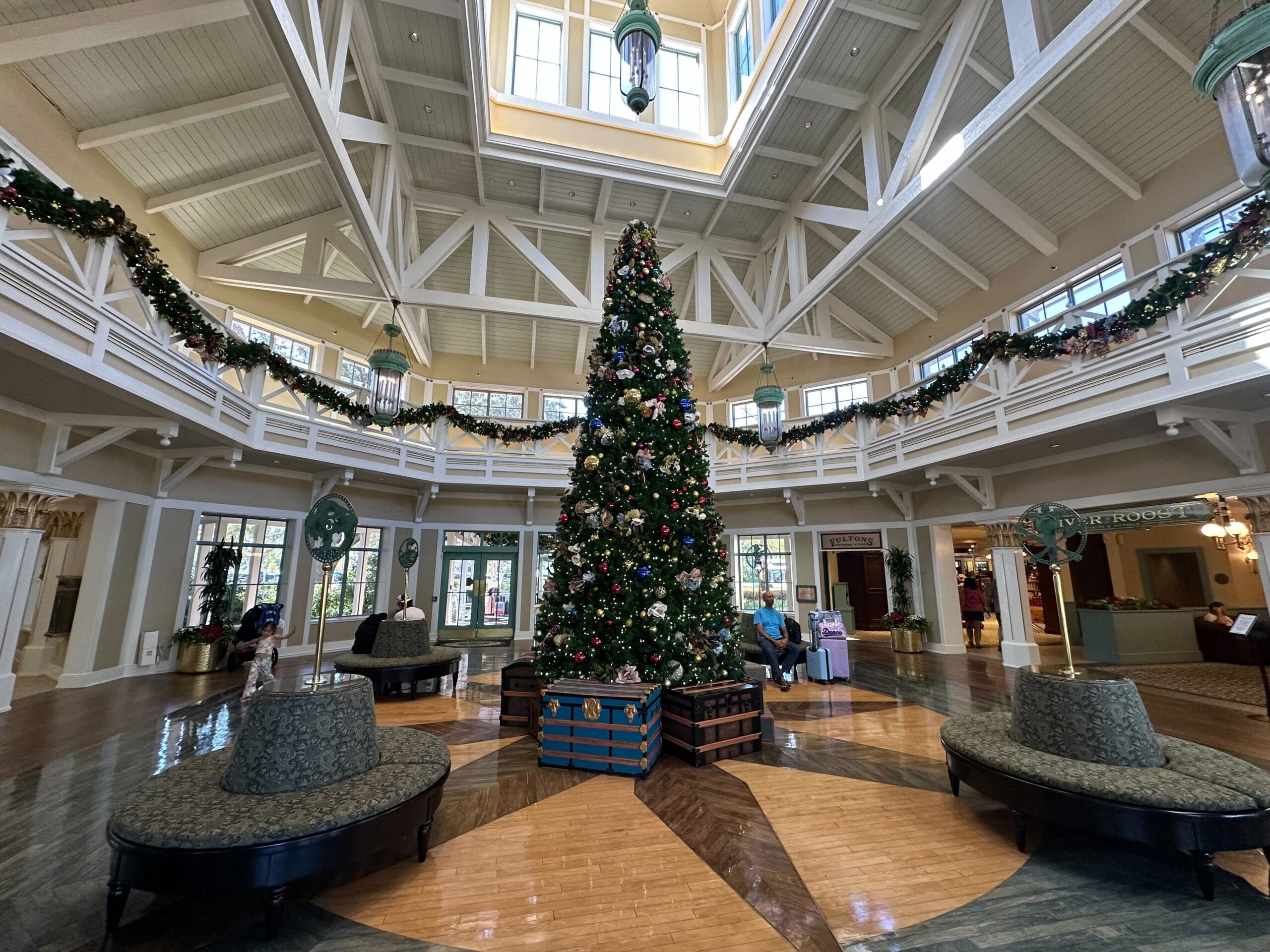 A large Christmas tree decorated with ornaments stands in the center of a spacious lobby. The room has festive garlands, high ceilings, and natural light from windows and skylights.