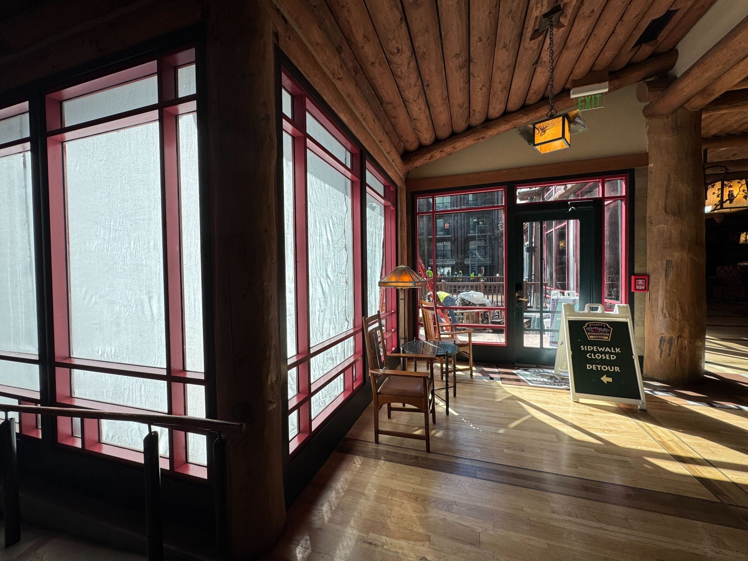 Sunlit lobby with wooden ceiling, covered windows, two chairs, a lamp, and a “Sidewalk Closed Detour” sign by the door.