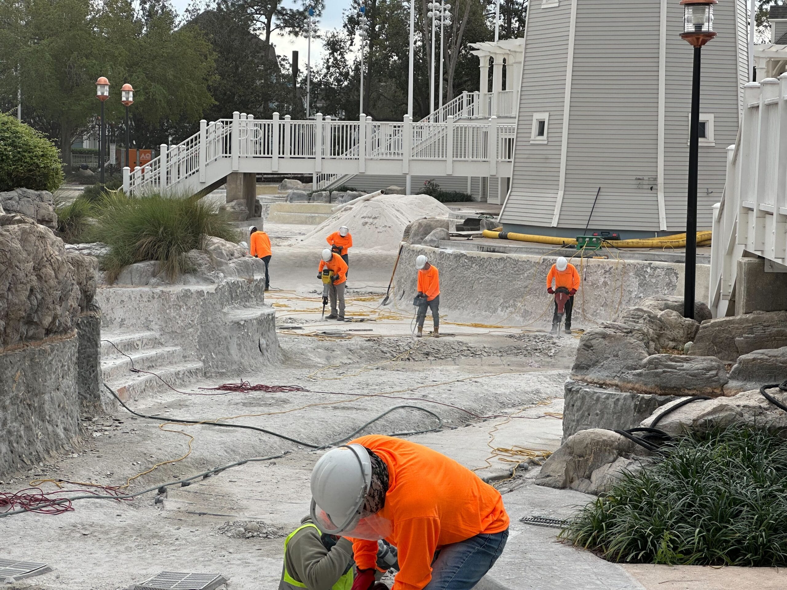 Workers in orange safety shirts and helmets conduct maintenance on an empty pool surrounded by rocks and structures.