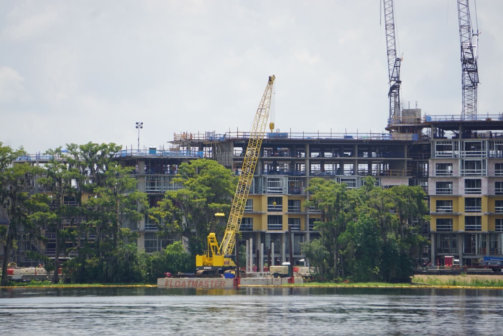 A yellow FLOATMASTER crane sits on a barge at a Lakeshore Lodge construction site by the lake.
