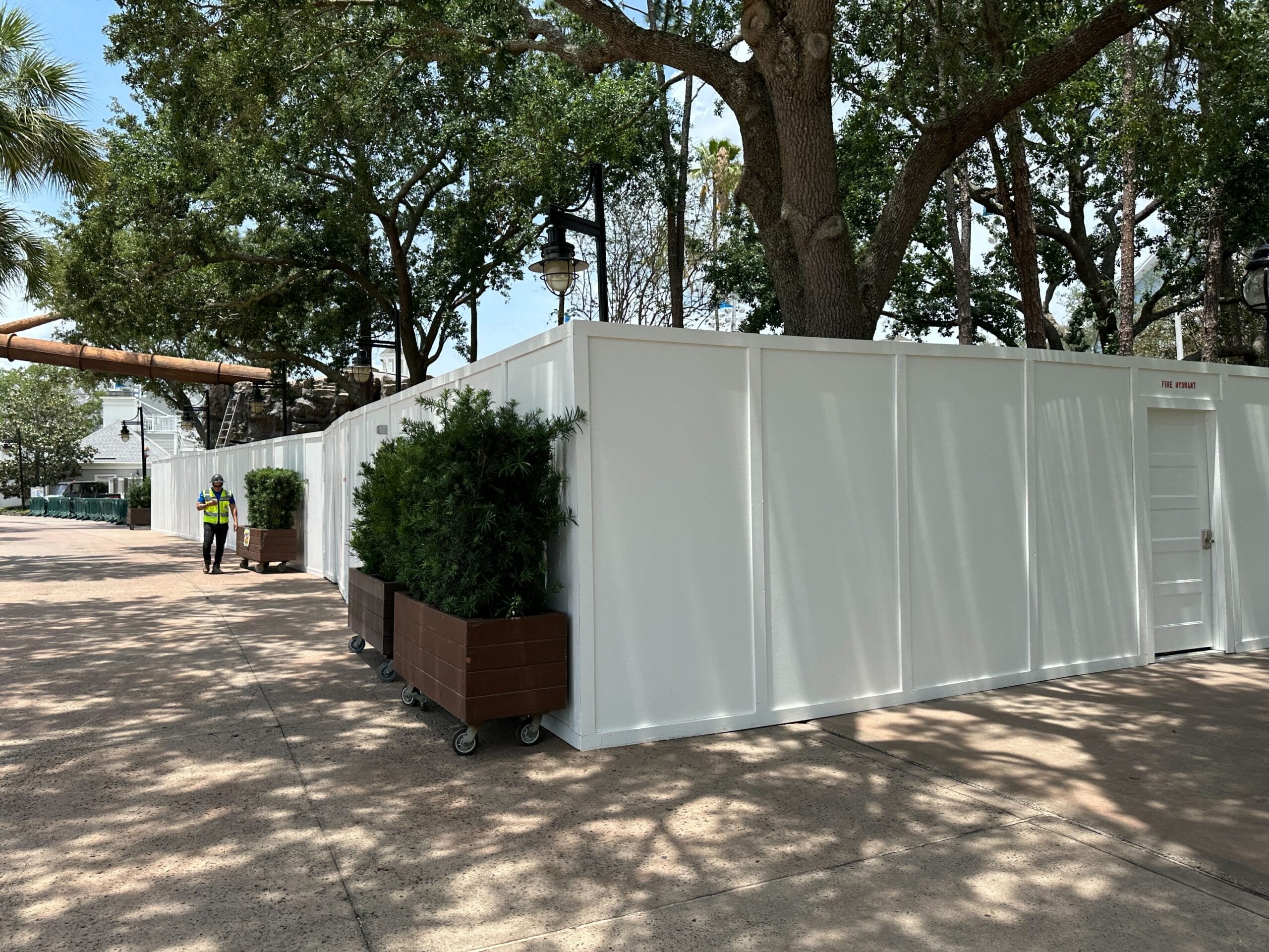 White construction walls with potted plants and trees, a worker in a safety vest stands nearby on a sunny day.