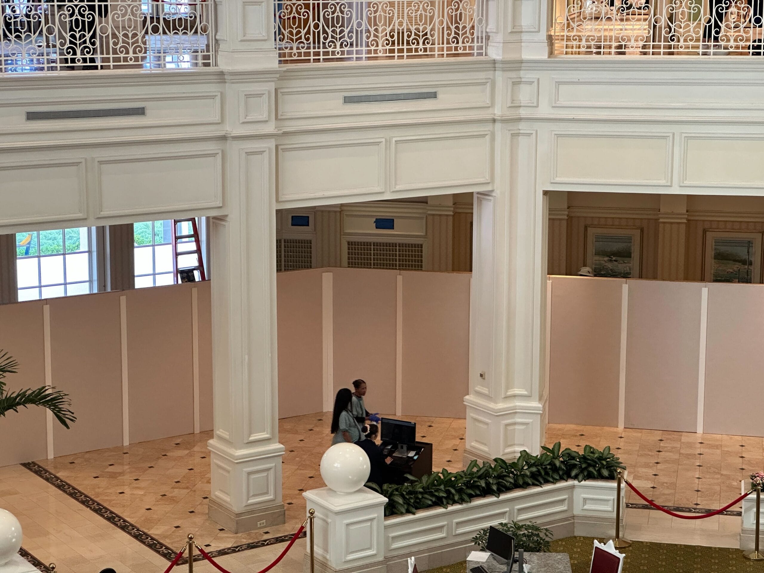 A person stands at a podium in a hotel lobby area, with white partition walls and decorative railings visible.