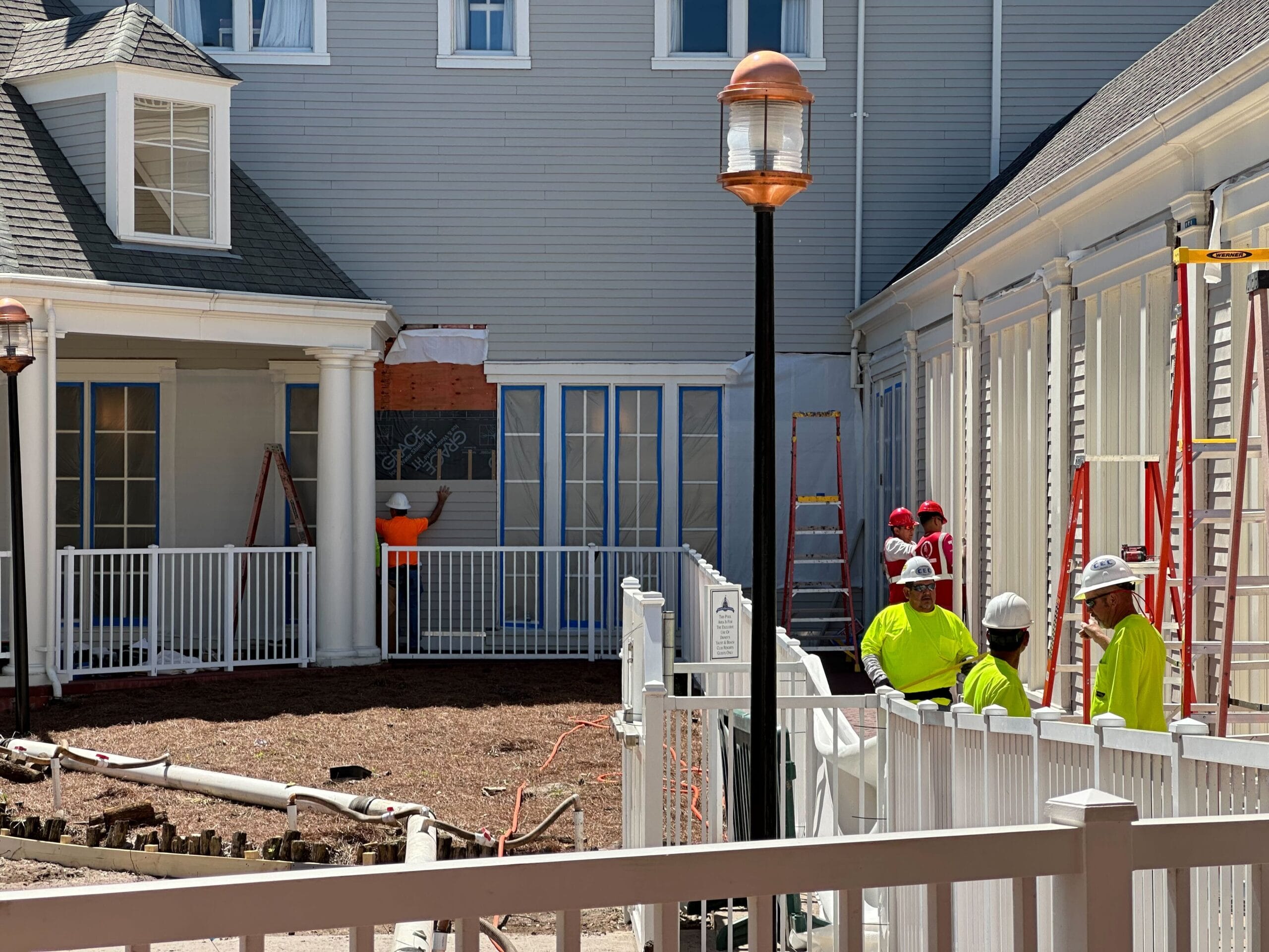 Construction workers in yellow vests and helmets were busy outside a building, surrounded by ladders and fencing. The scene unfolded under a clear sky near Disney’s Yacht Club, their work contributing to the magic of nearby attractions like Stormalong Bay Pool.
