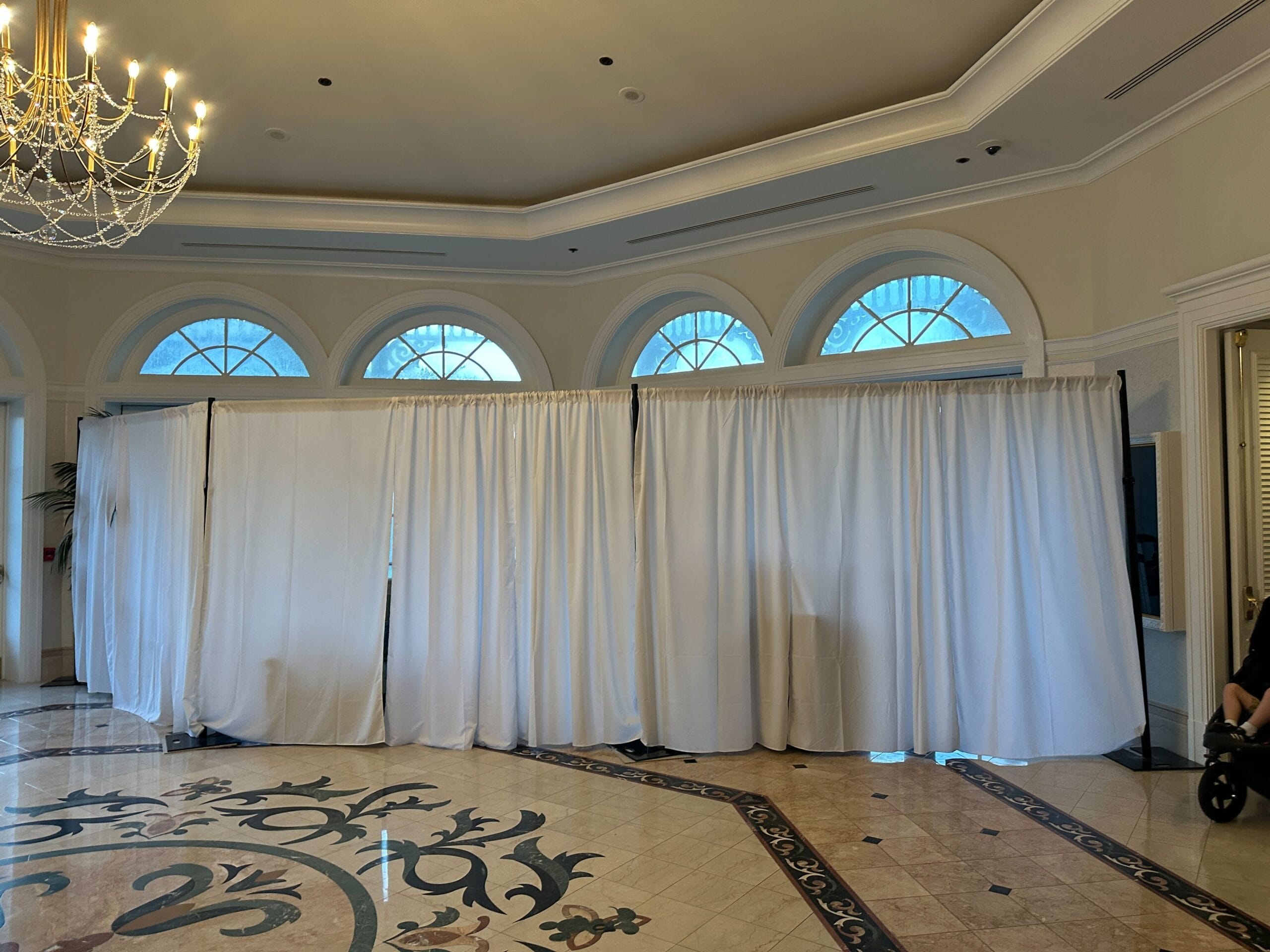 White curtains partition a large, elegant Grand Floridian room with arched windows, chandelier, and patterned marble floor.