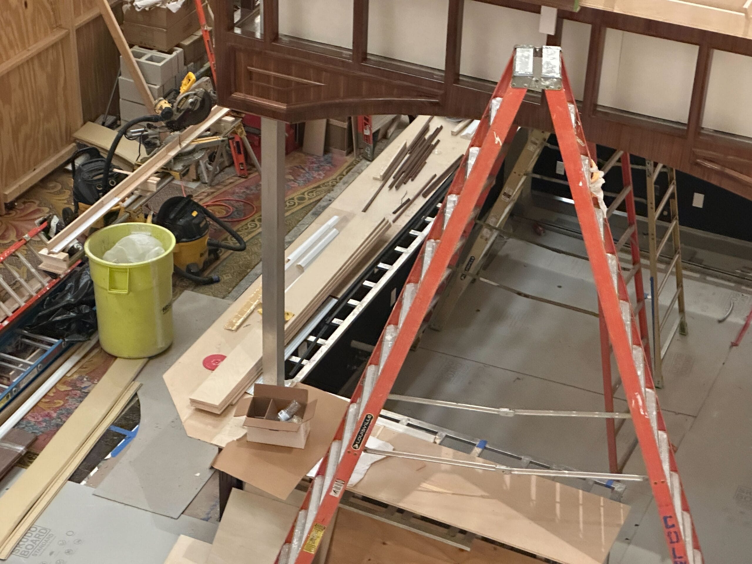 Construction site with a ladder, wood planks, tools, a yellow trash bin, and scattered building materials.