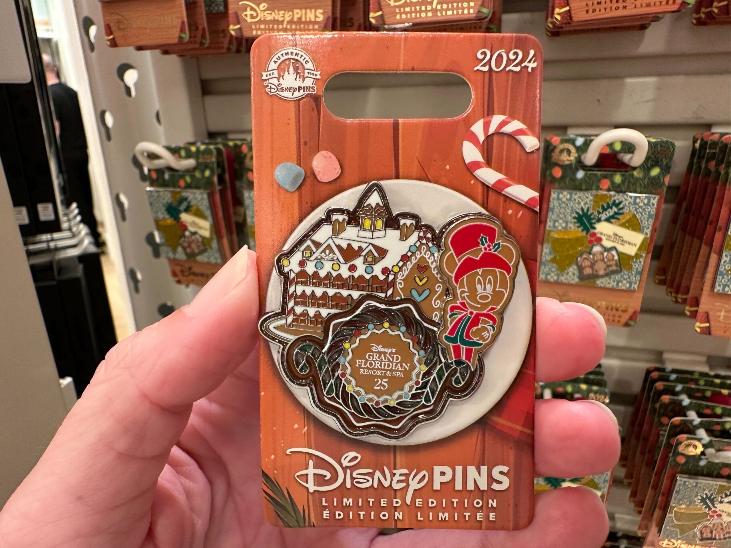 Hand holding a 2024 limited edition Disney pin featuring a gingerbread house, peppermint stick, and festive character in uniform, displayed in packaging.
