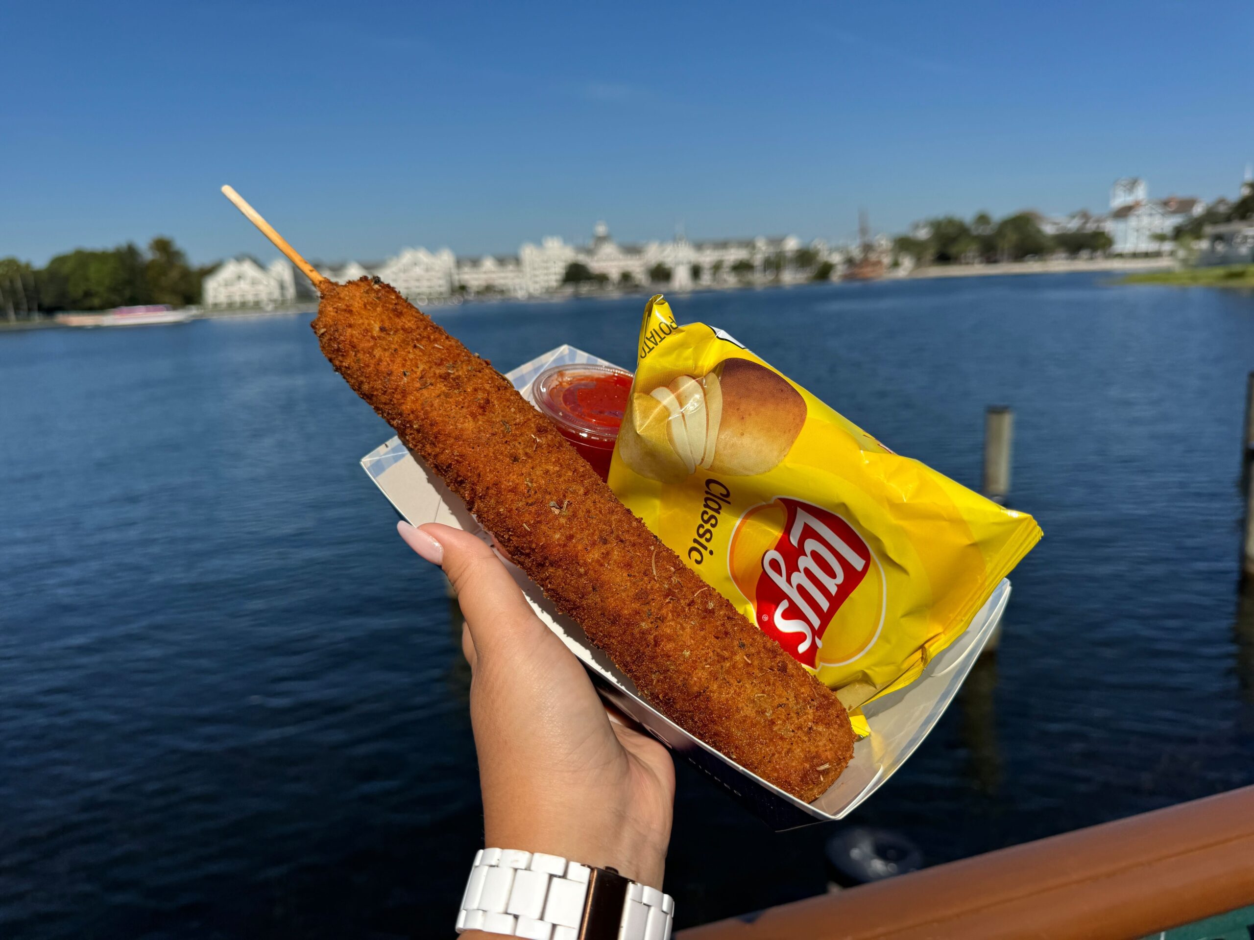 A hand holds a deep-fried corn dog on a stick and a bag of Lay's Classic chips against a scenic waterfront backdrop. A hand holds a deep-fried corn dog on a stick and a bag of Lay's Classic chips against a scenic waterfront backdrop.