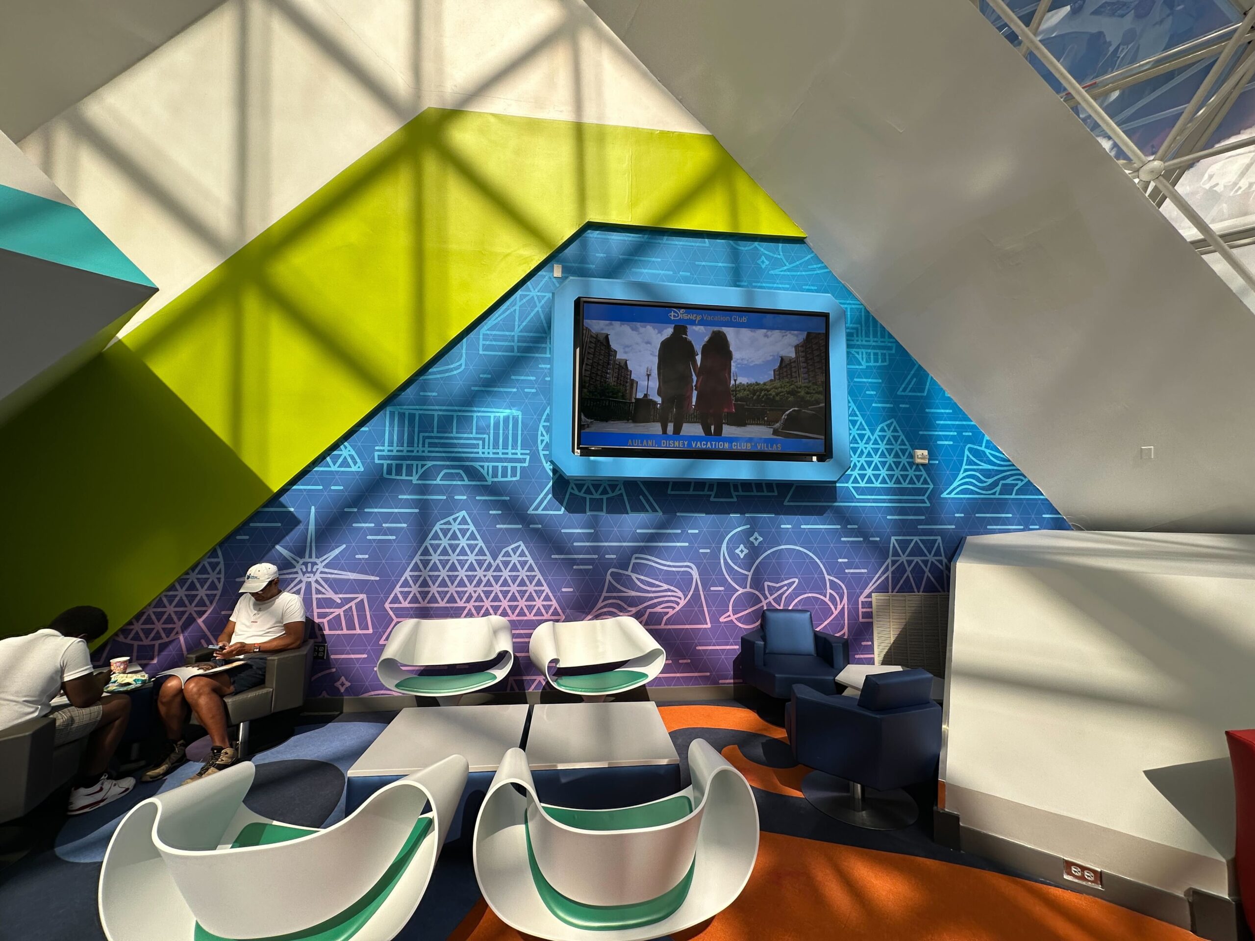 Colorful lounge with geometric wall designs, a TV screen displaying a show, and empty modern chairs. Two people sit on the left, one wearing a white cap. Sunlight casts shadows on the wall. Colorful lounge with geometric wall designs, a TV screen displaying a show, and empty modern chairs. Two people sit on the left, one wearing a white cap. Sunlight casts shadows on the wall.