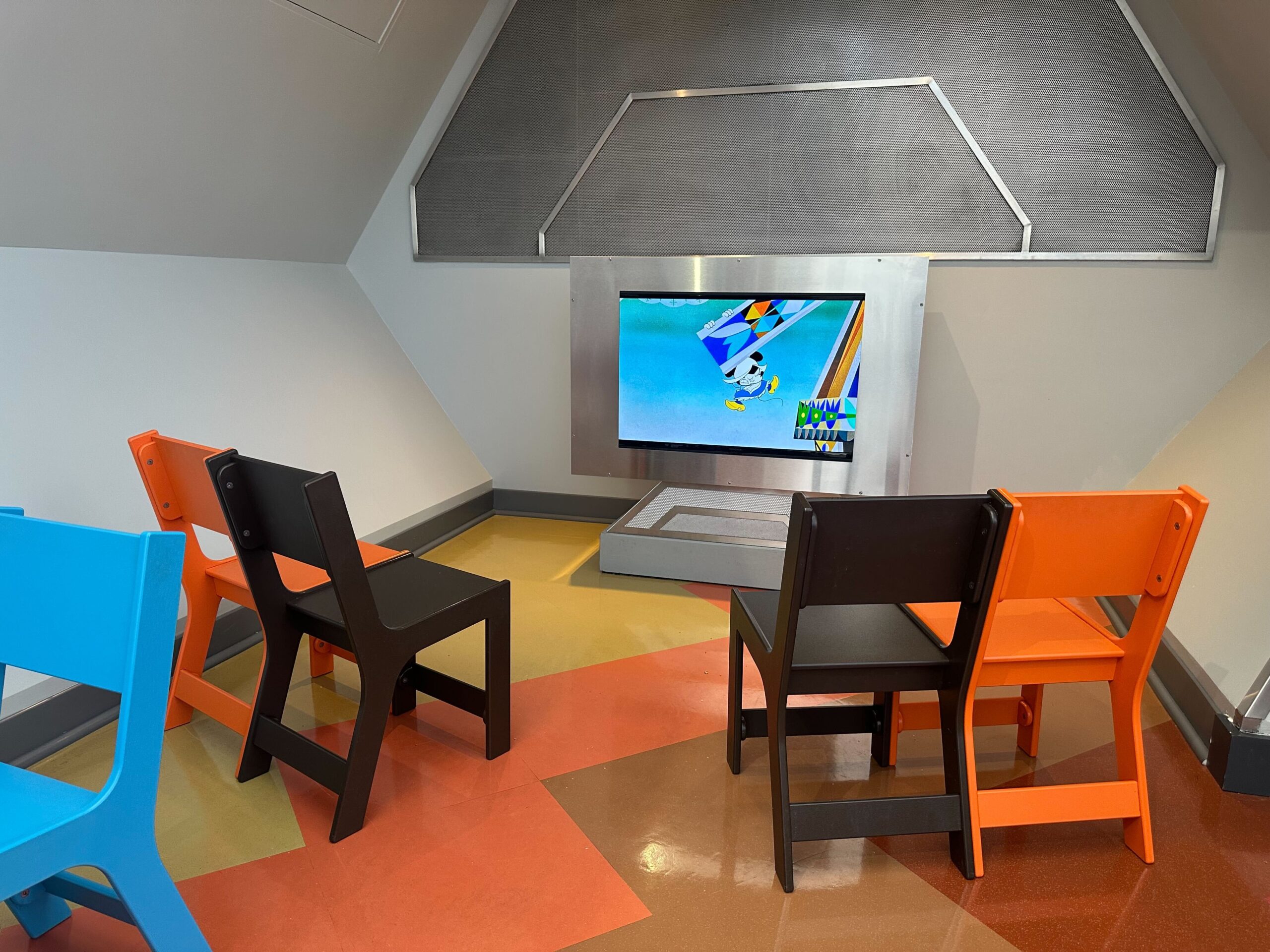 A room with colorful chairs facing a TV screen displaying an animated show. The floor has a geometric pattern in orange, yellow, and brown. A room with colorful chairs facing a TV screen displaying an animated show. The floor has a geometric pattern in orange, yellow, and brown.