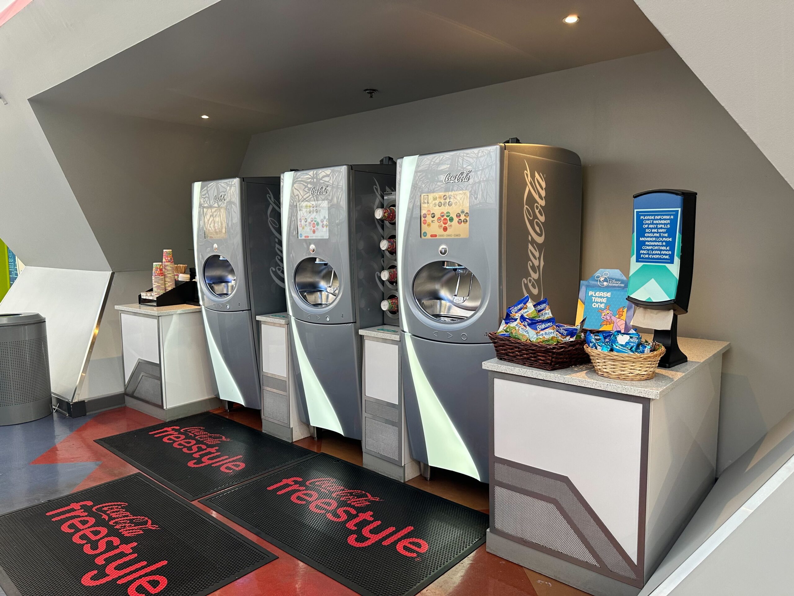 A row of four Coca-Cola Freestyle machines with a display of cups and a basket of snacks on the counter. A row of four Coca-Cola Freestyle machines with a display of cups and a basket of snacks on the counter.
