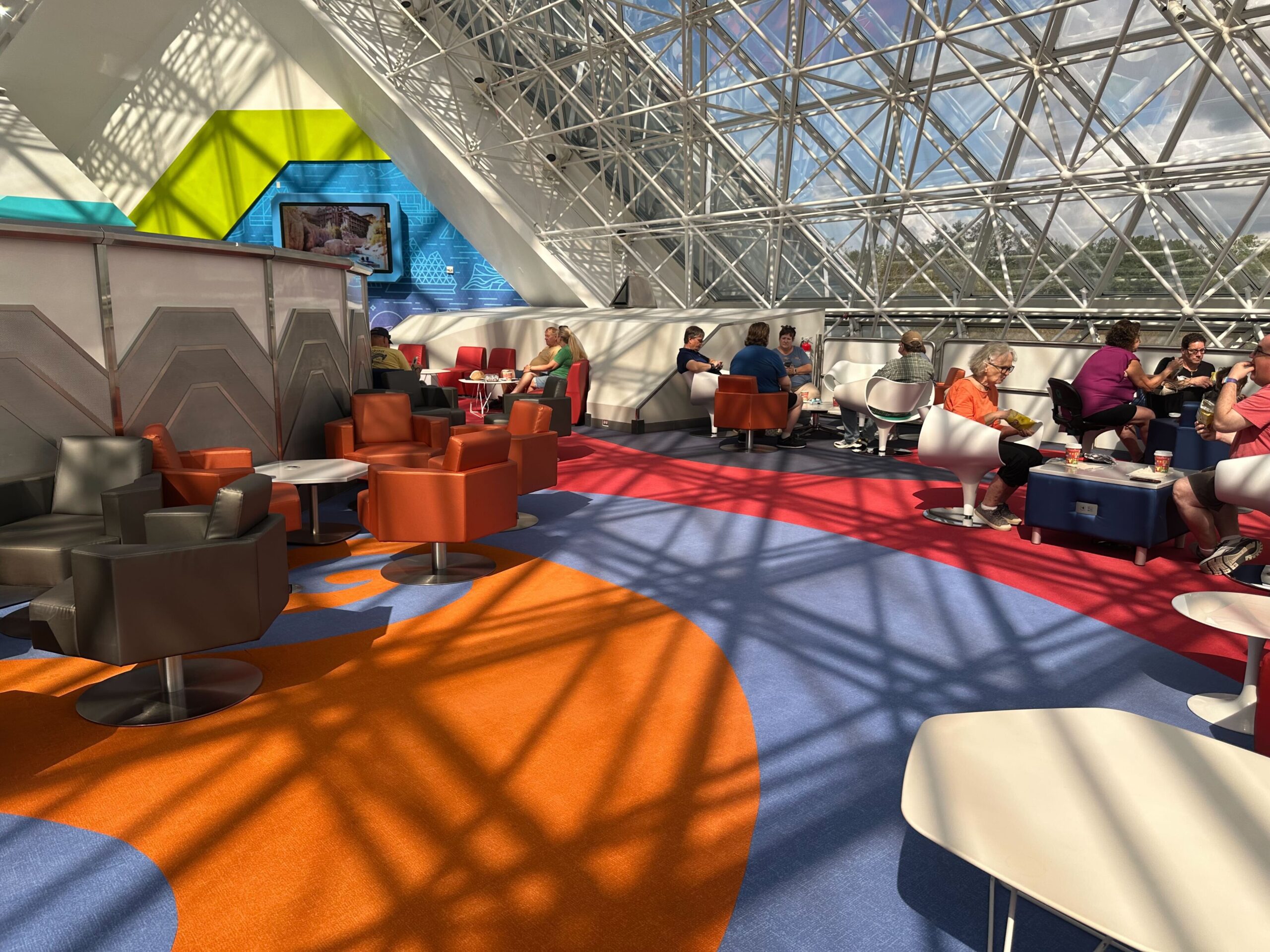 People sitting in a brightly lit, modern lounge area with colorful chairs and geometric designs on the walls and carpet. People sitting in a brightly lit, modern lounge area with colorful chairs and geometric designs on the walls and carpet.