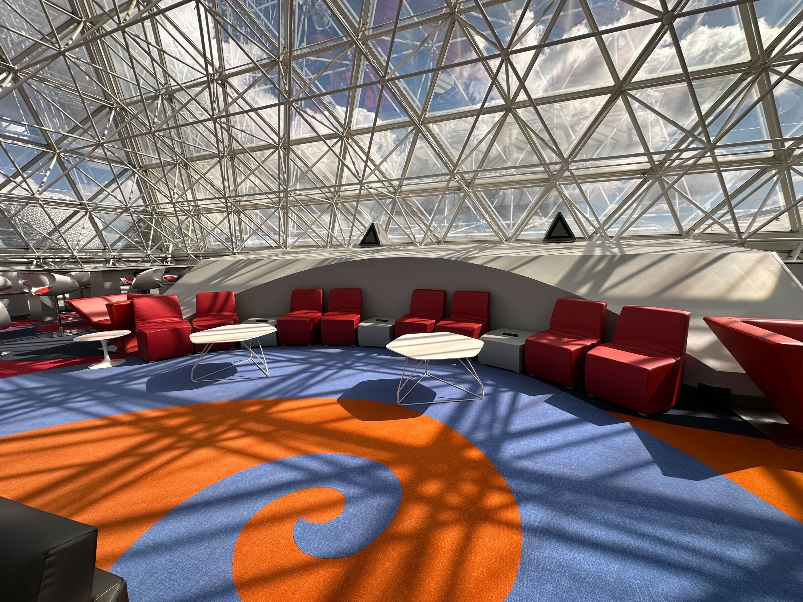 A brightly lit lounge with red chairs and round white tables, set against a geometric glass ceiling. The carpet features orange and blue swirls. A brightly lit lounge with red chairs and round white tables, set against a geometric glass ceiling. The carpet features orange and blue swirls.