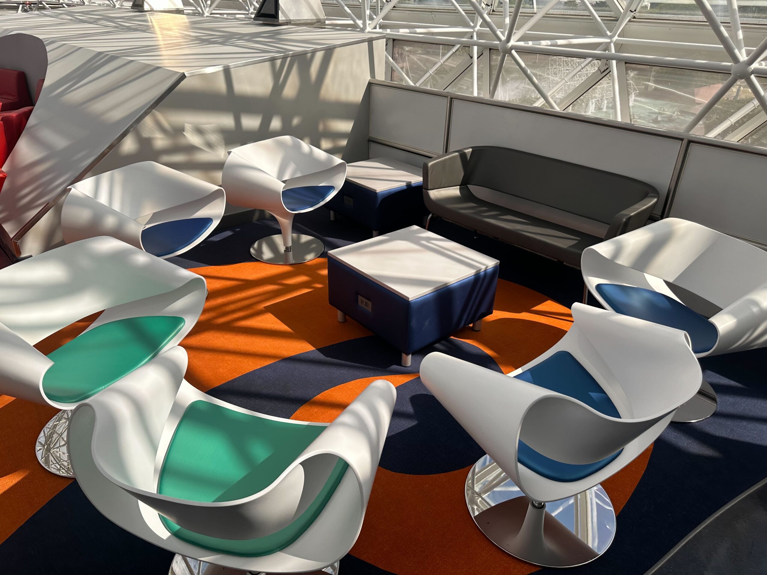 A seating area with modern white chairs and a gray sofa is arranged around square tables on an orange and blue carpet under a geometric glass roof. A seating area with modern white chairs and a gray sofa is arranged around square tables on an orange and blue carpet under a geometric glass roof.
