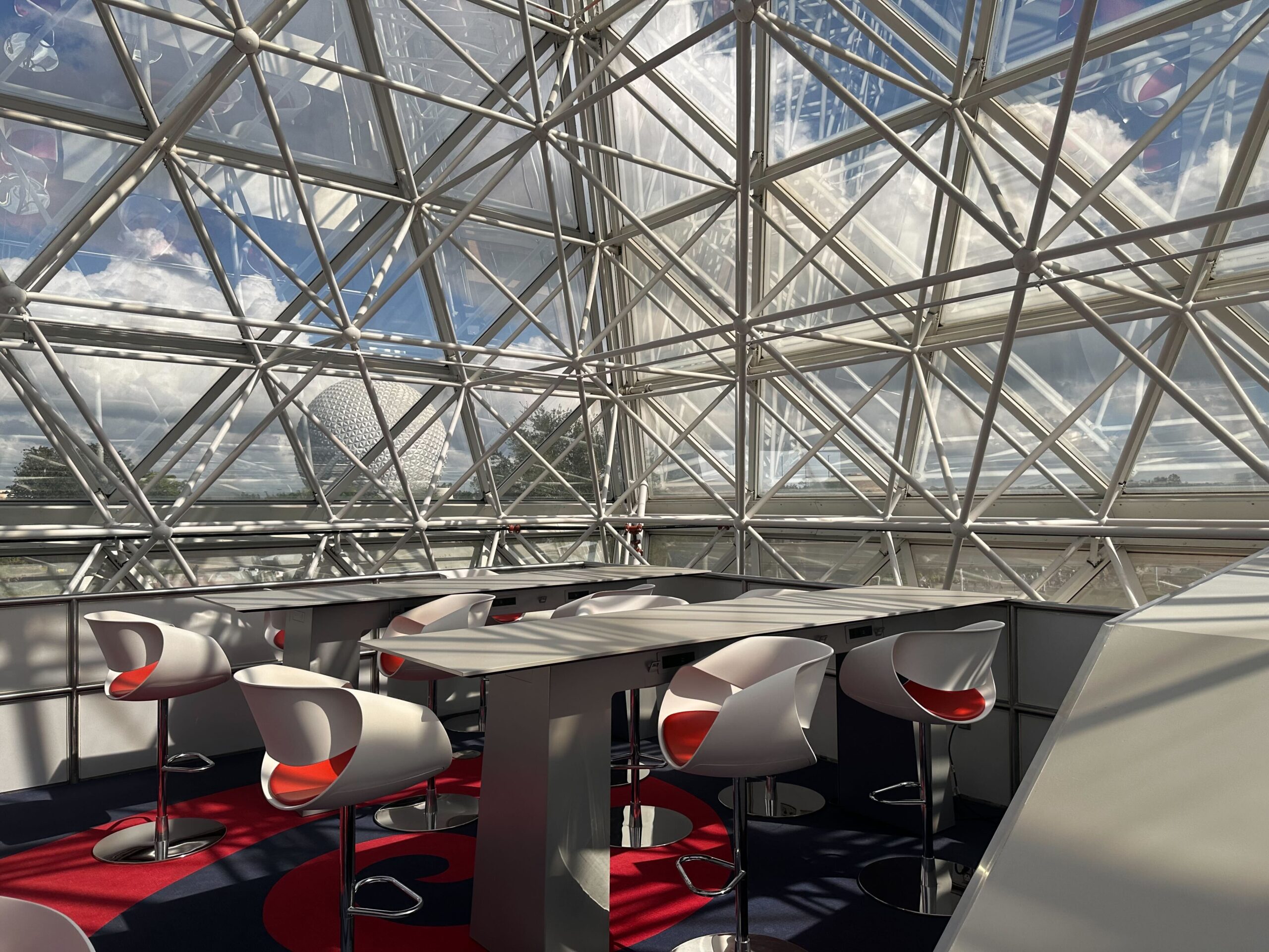 A modern glass-walled meeting room with white chairs around a table, featuring a geometric ceiling structure and a view of partly cloudy skies. A modern glass-walled meeting room with white chairs around a table, featuring a geometric ceiling structure and a view of partly cloudy skies.