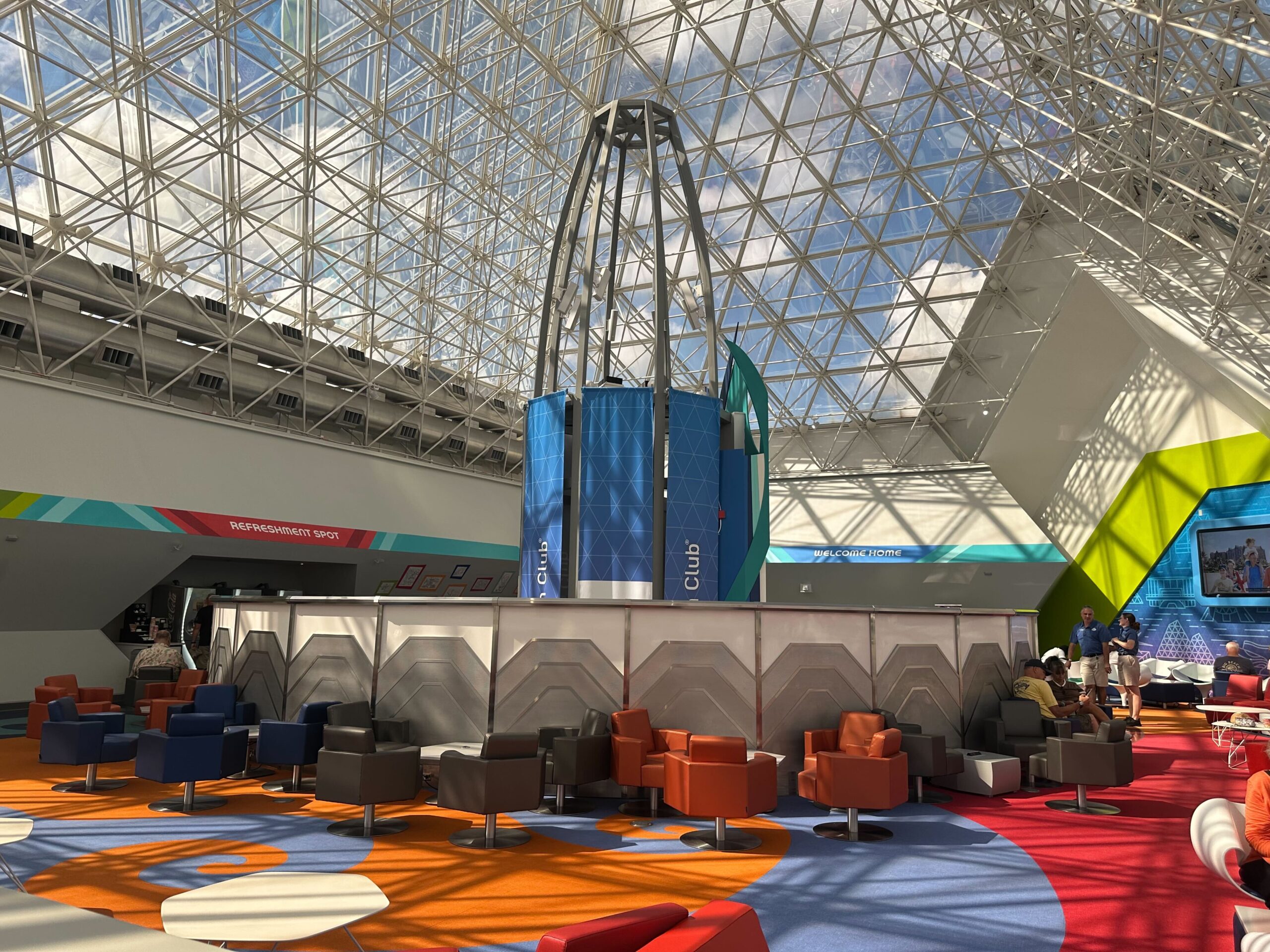 Futuristic lounge interior with colorful chairs, under a large glass ceiling and geometric design elements. A cylindrical structure is in the center. Futuristic lounge interior with colorful chairs, under a large glass ceiling and geometric design elements. A cylindrical structure is in the center.
