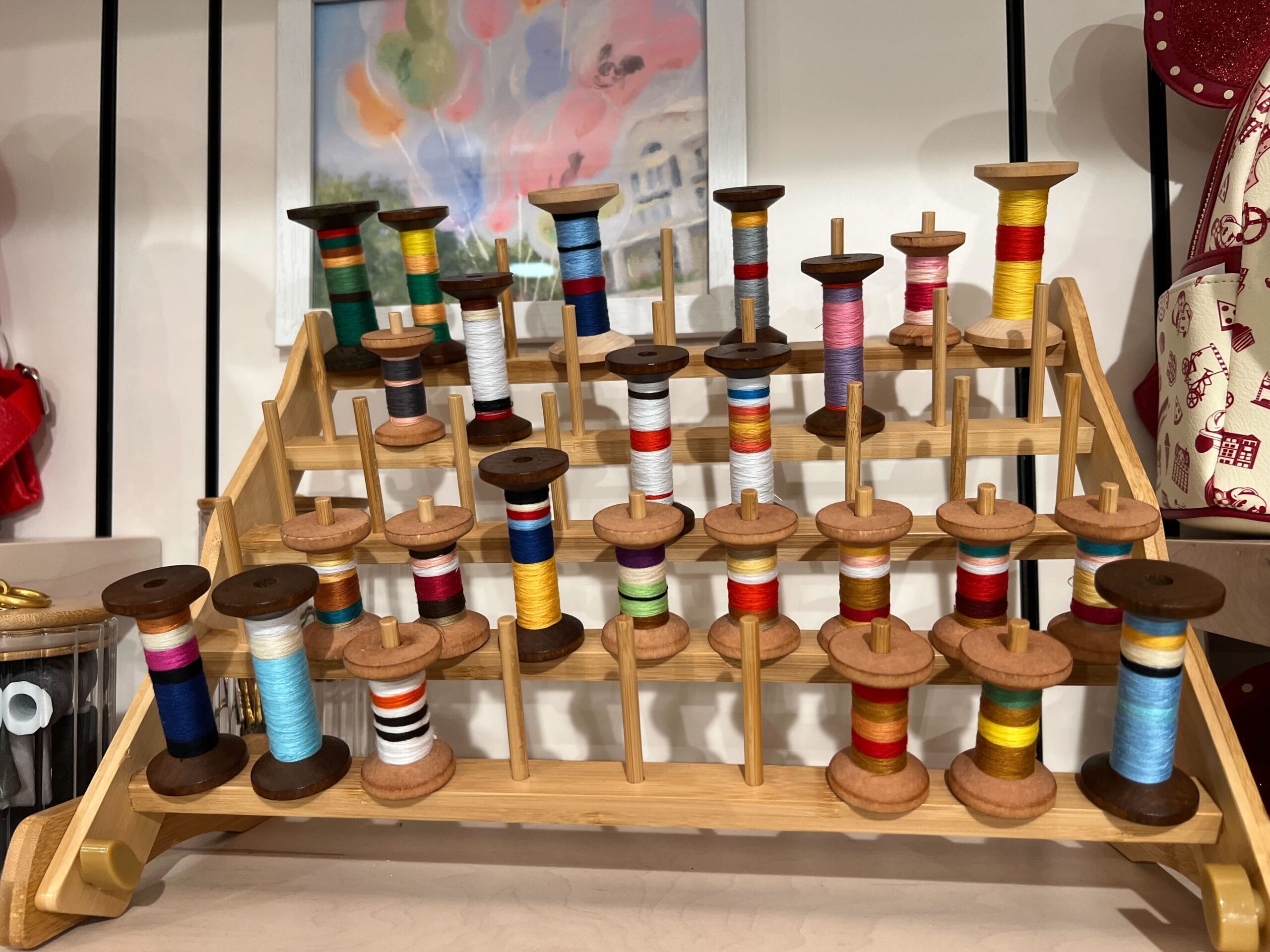 A wooden rack at Thimbles & Threads holds colorful spools of thread, arranged in rows, with a painting in the background. A wooden rack at Thimbles & Threads holds colorful spools of thread, arranged in rows, with a painting in the background.