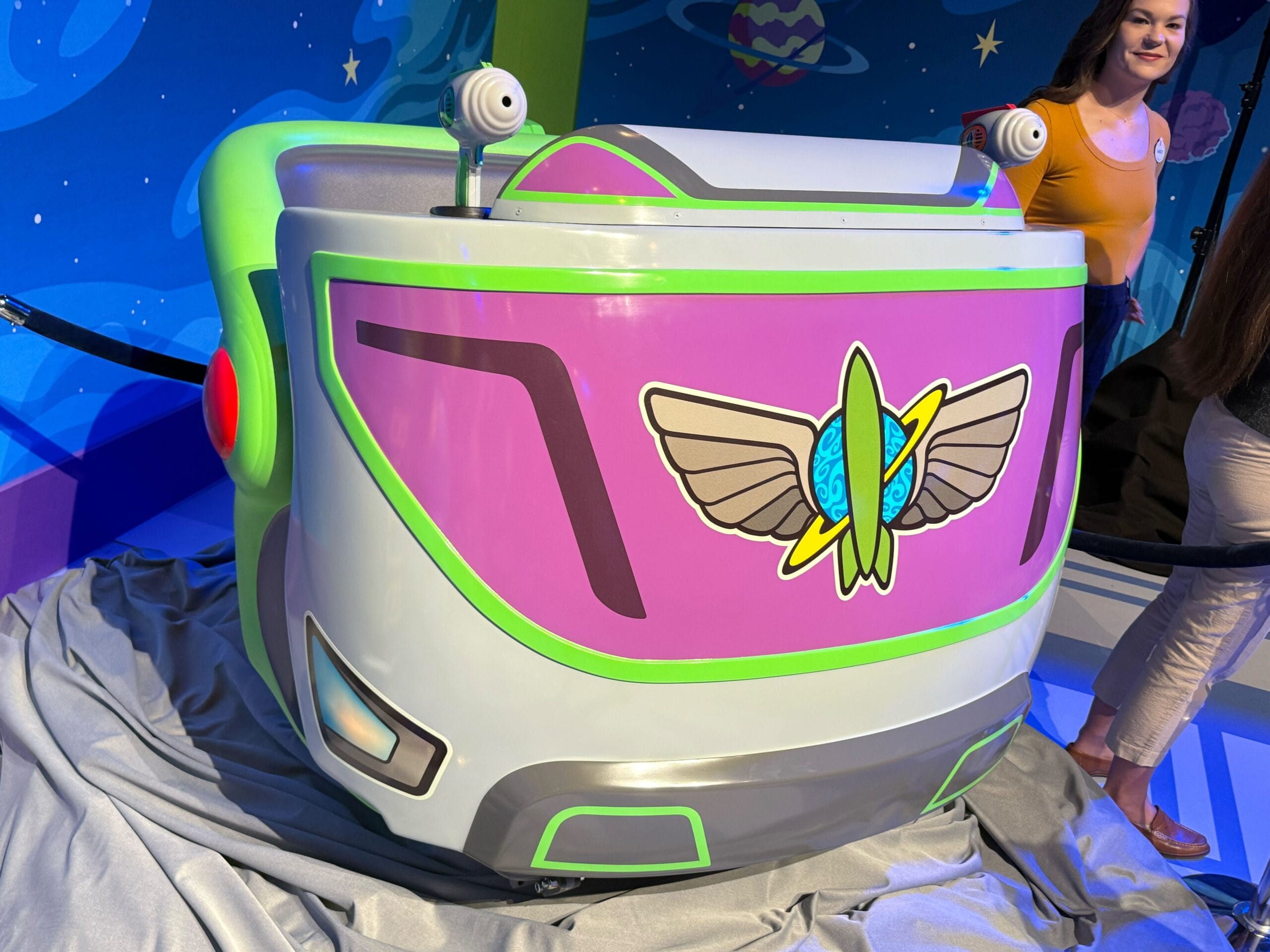 First look: Colorful Buzz Lightyear Space Ranger Spin ride vehicle with wings logo, indoors with two people nearby. First look: Colorful Buzz Lightyear Space Ranger Spin ride vehicle with wings logo, indoors with two people nearby.