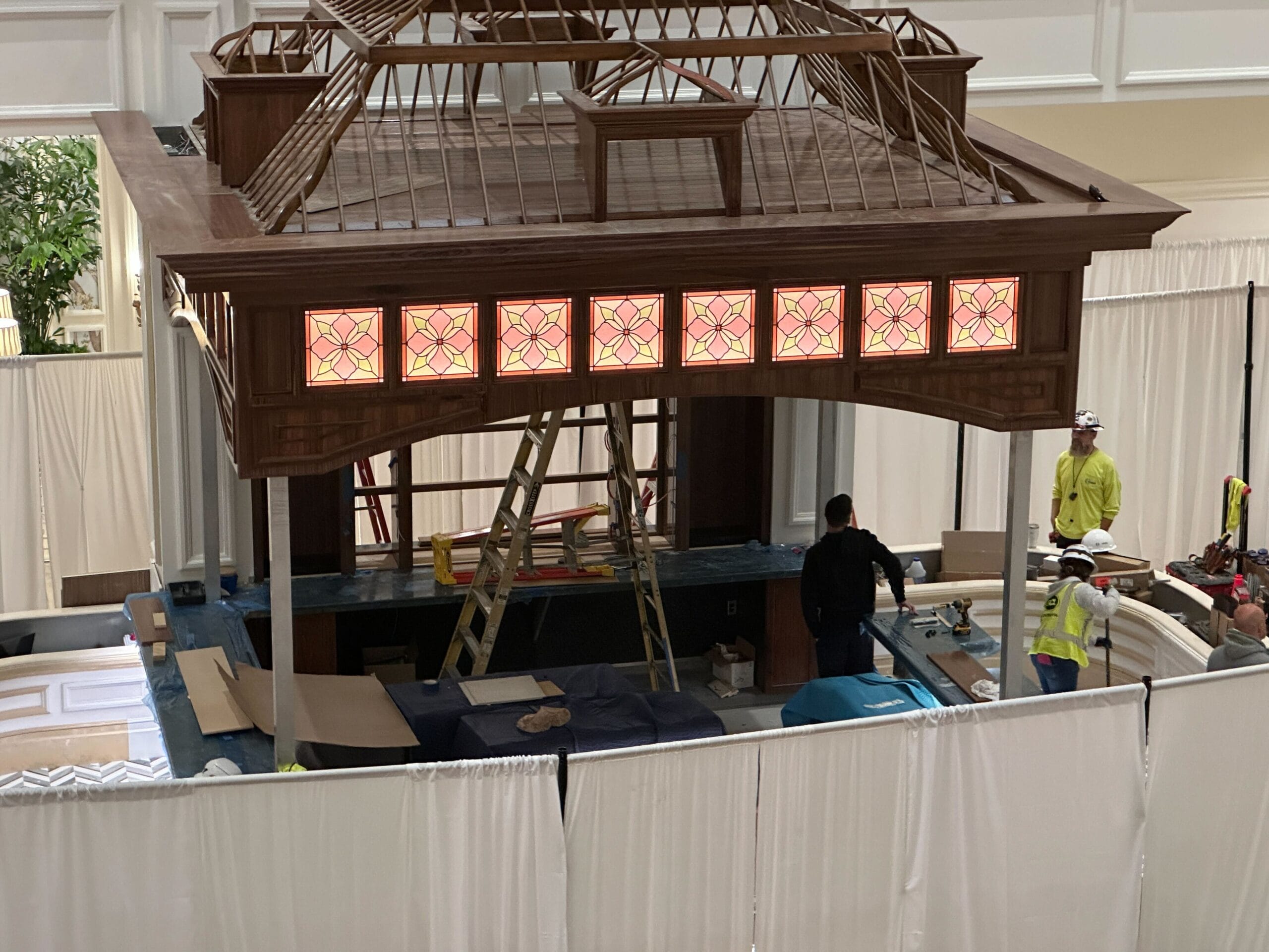 Workers build a wooden structure with stained-glass panels at the Birdcage Bar, similar to themed Disney park interiors, white curtains nearby. Workers build a wooden structure with stained-glass panels at the Birdcage Bar, similar to themed Disney park interiors, white curtains nearby.