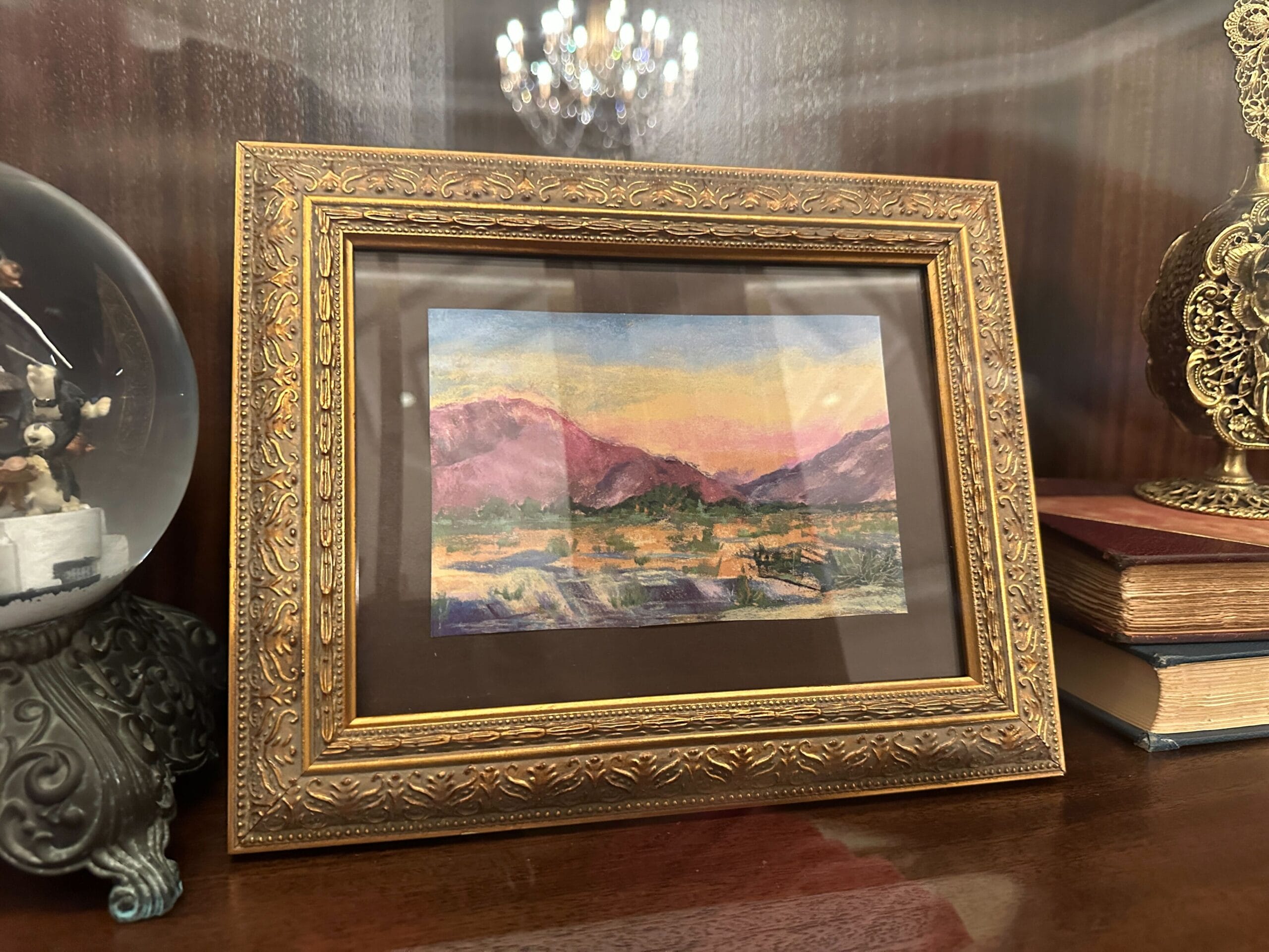 A framed painting of mountains and a sunset with warm lighting, on a wooden shelf in a glass-front display case in the refurbished Grand Floridian lobby bar. A framed painting of mountains and a sunset with warm lighting, on a wooden shelf in a glass-front display case in the refurbished Grand Floridian lobby bar.