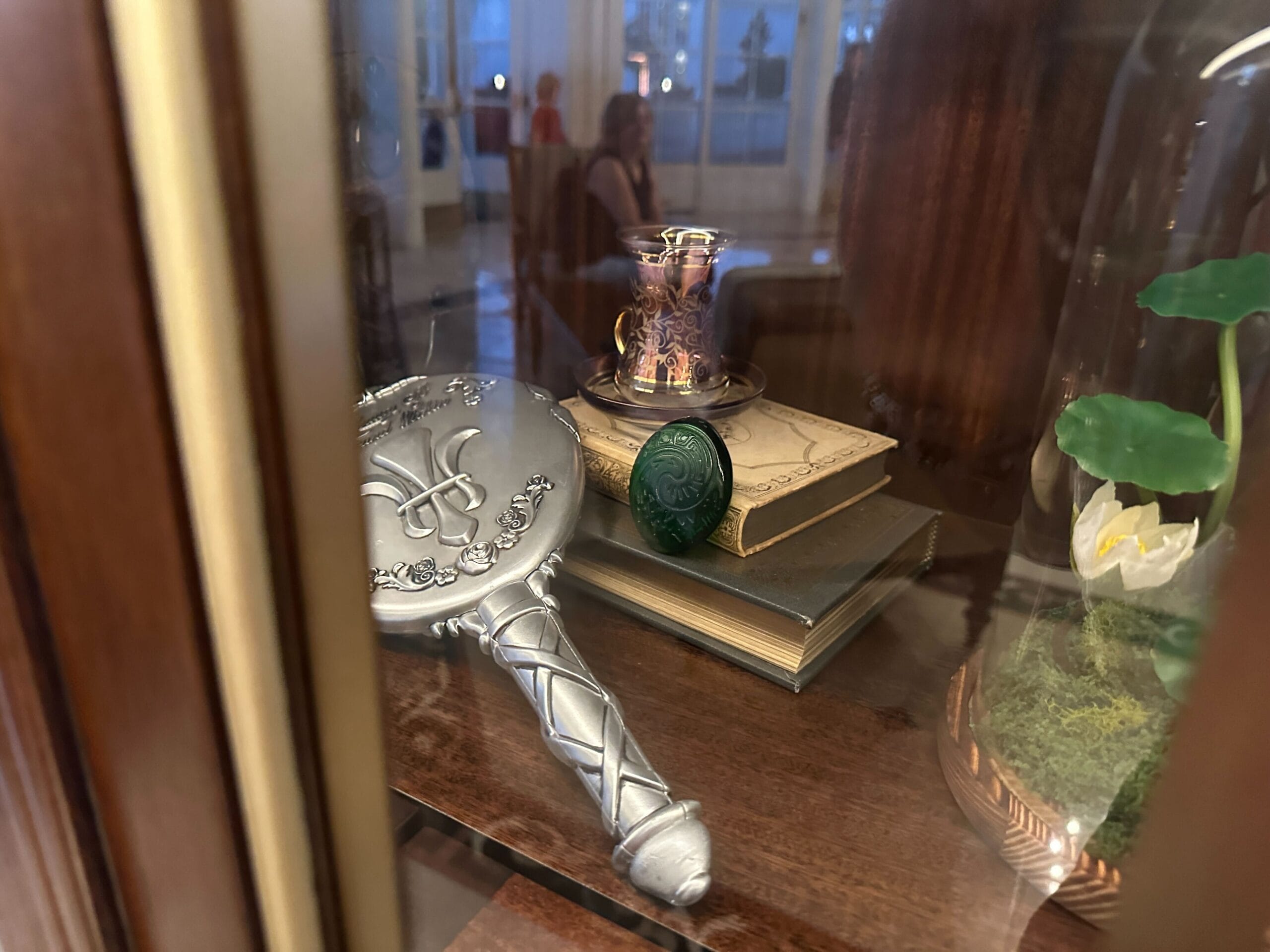 A silver hand mirror, green stone, glass teacup, and bell-jarred flower in a glass-front display case at the refurbished Grand Floridian lobby bar. A silver hand mirror, green stone, glass teacup, and bell-jarred flower in a glass-front display case at the refurbished Grand Floridian lobby bar.