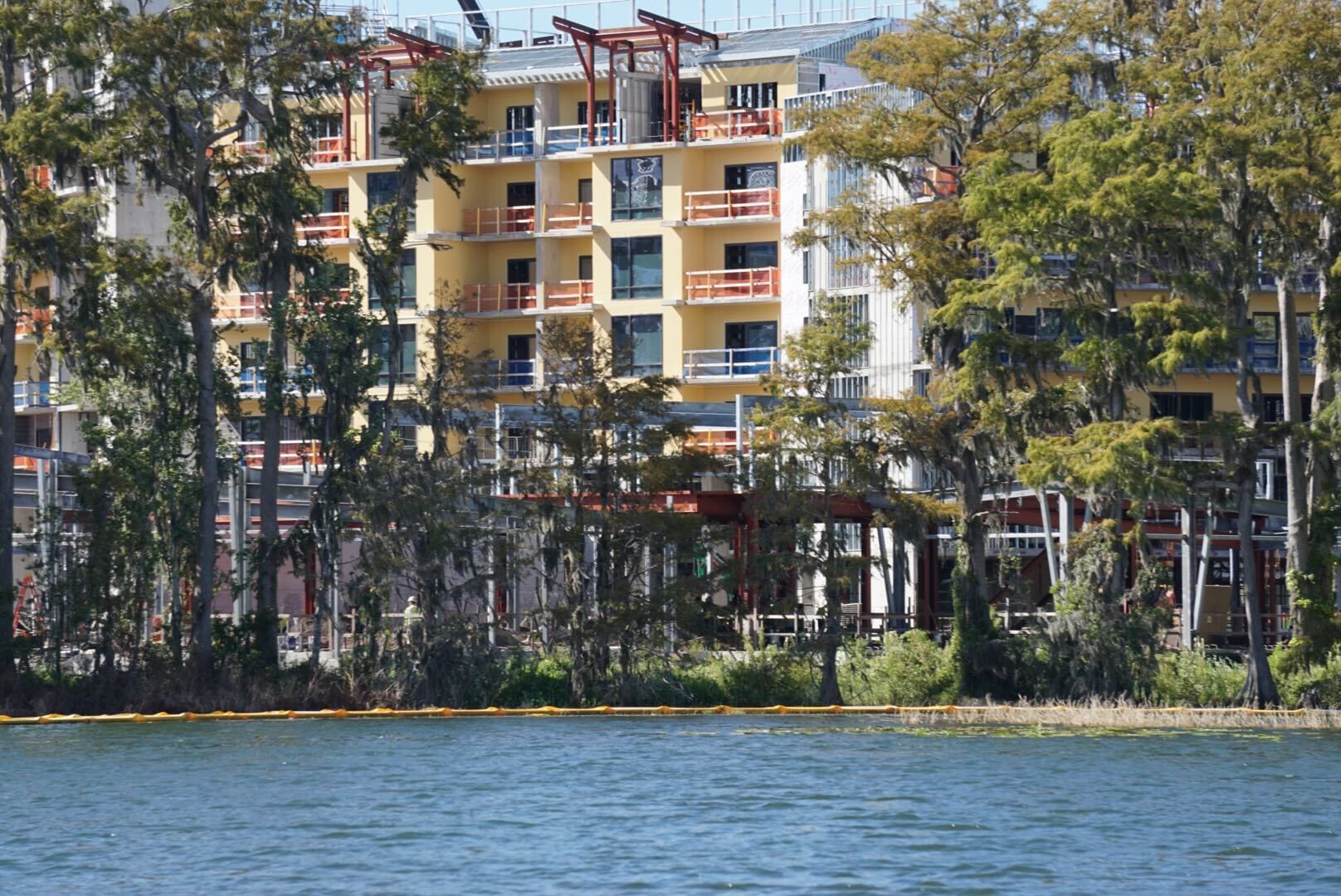 A yellow building under construction, reminiscent of Epcot's ongoing projects, rises behind trees by a blue lakeshore. A yellow building under construction, reminiscent of Epcot's ongoing projects, rises behind trees by a blue lakeshore.