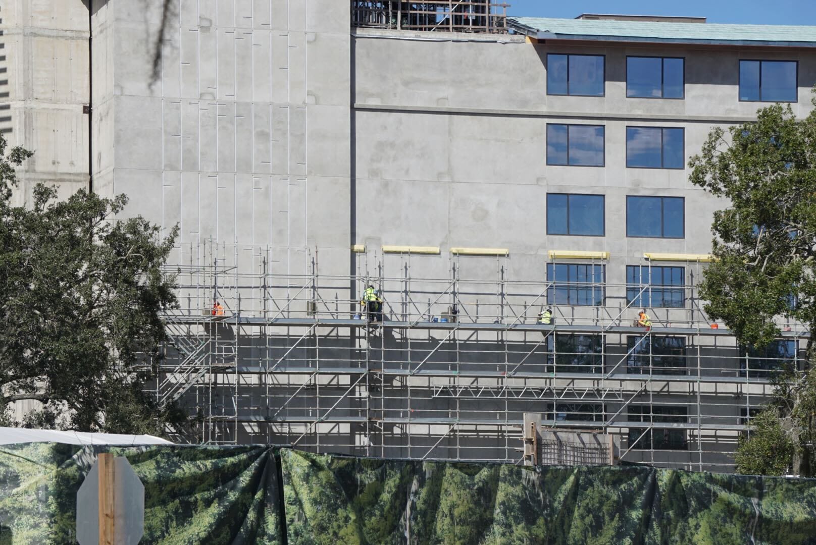 Construction workers on scaffolding at an unfinished building—Lakeshore update with green fencing and trees, similar to EPCOT projects. Construction workers on scaffolding at an unfinished building—Lakeshore update with green fencing and trees, similar to EPCOT projects.