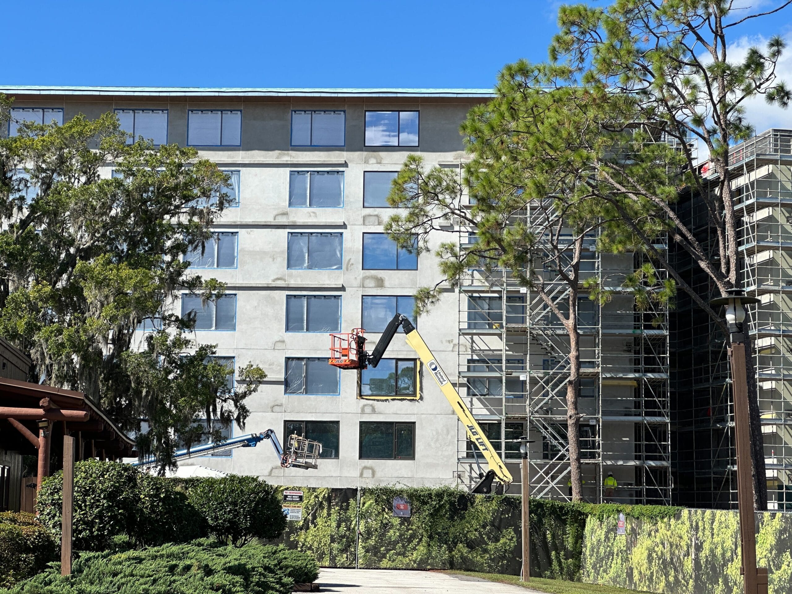 Two construction lifts repair a multi-story building’s facade, surrounded by trees and scaffolding in a Disney park update scene. Two construction lifts repair a multi-story building’s facade, surrounded by trees and scaffolding in a Disney park update scene.