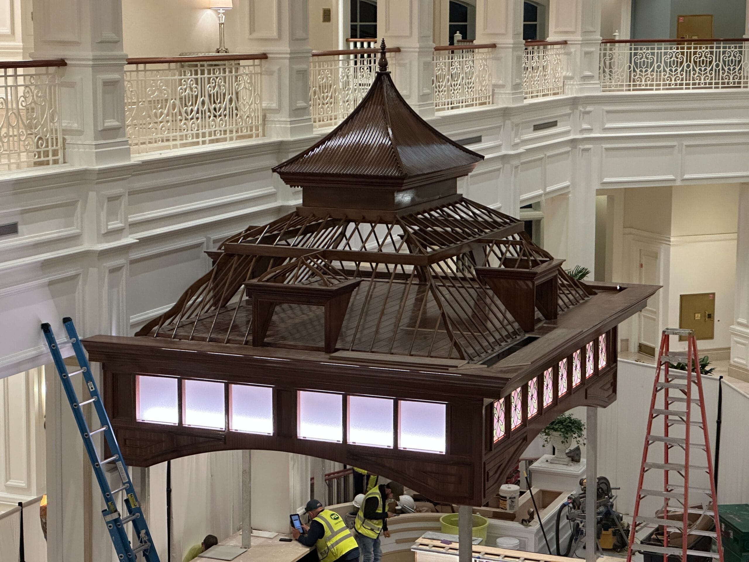 Two workers assemble a detailed wooden gazebo-like structure inside a grand, bright venue reminiscent of Disney park architecture. Two workers assemble a detailed wooden gazebo-like structure inside a grand, bright venue reminiscent of Disney park architecture.