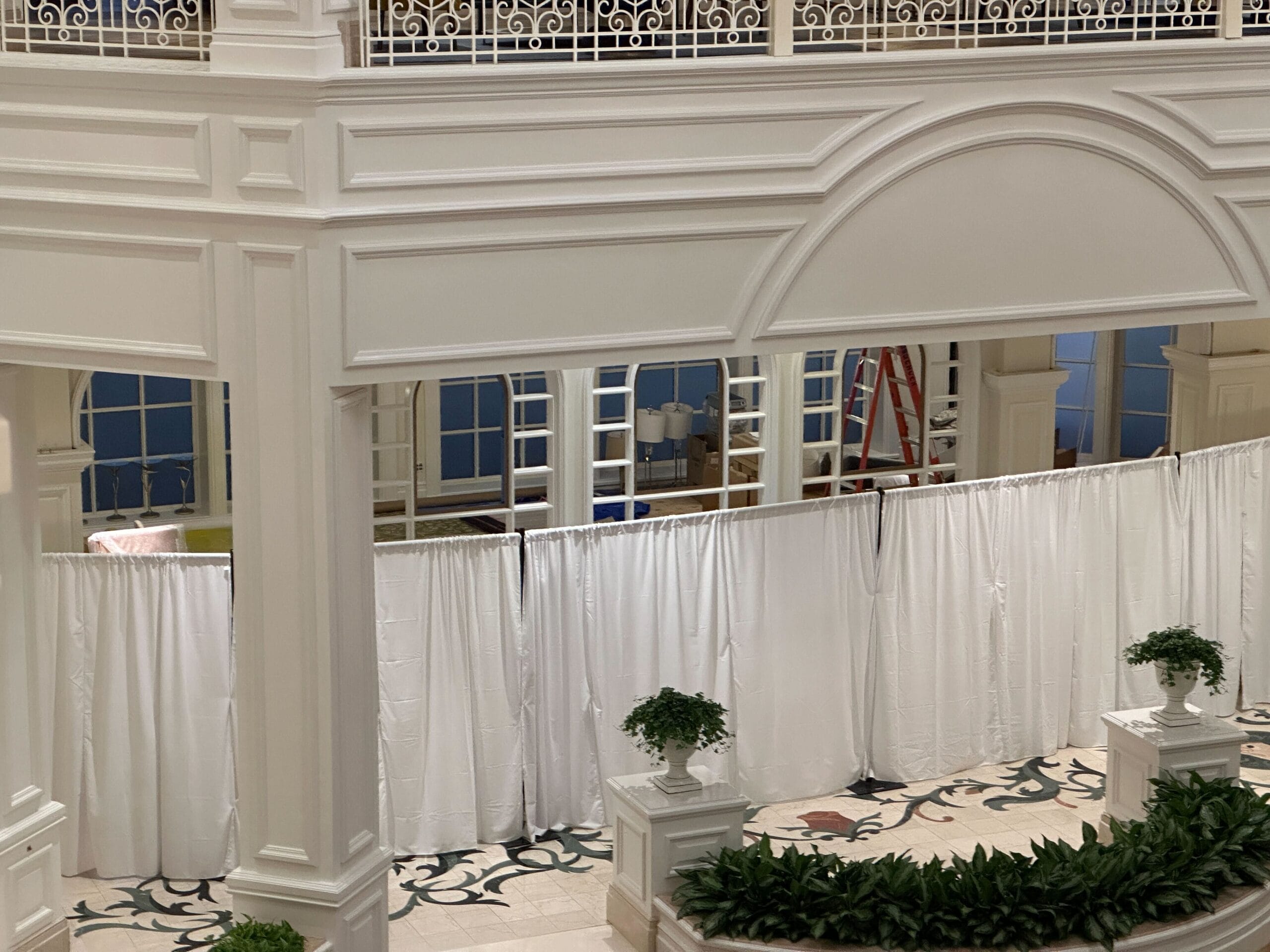 White curtains hide part of a grand hotel lobby near Disney, with a red ladder and supplies peeking out like backstage magic. White curtains hide part of a grand hotel lobby near Disney, with a red ladder and supplies peeking out like backstage magic.