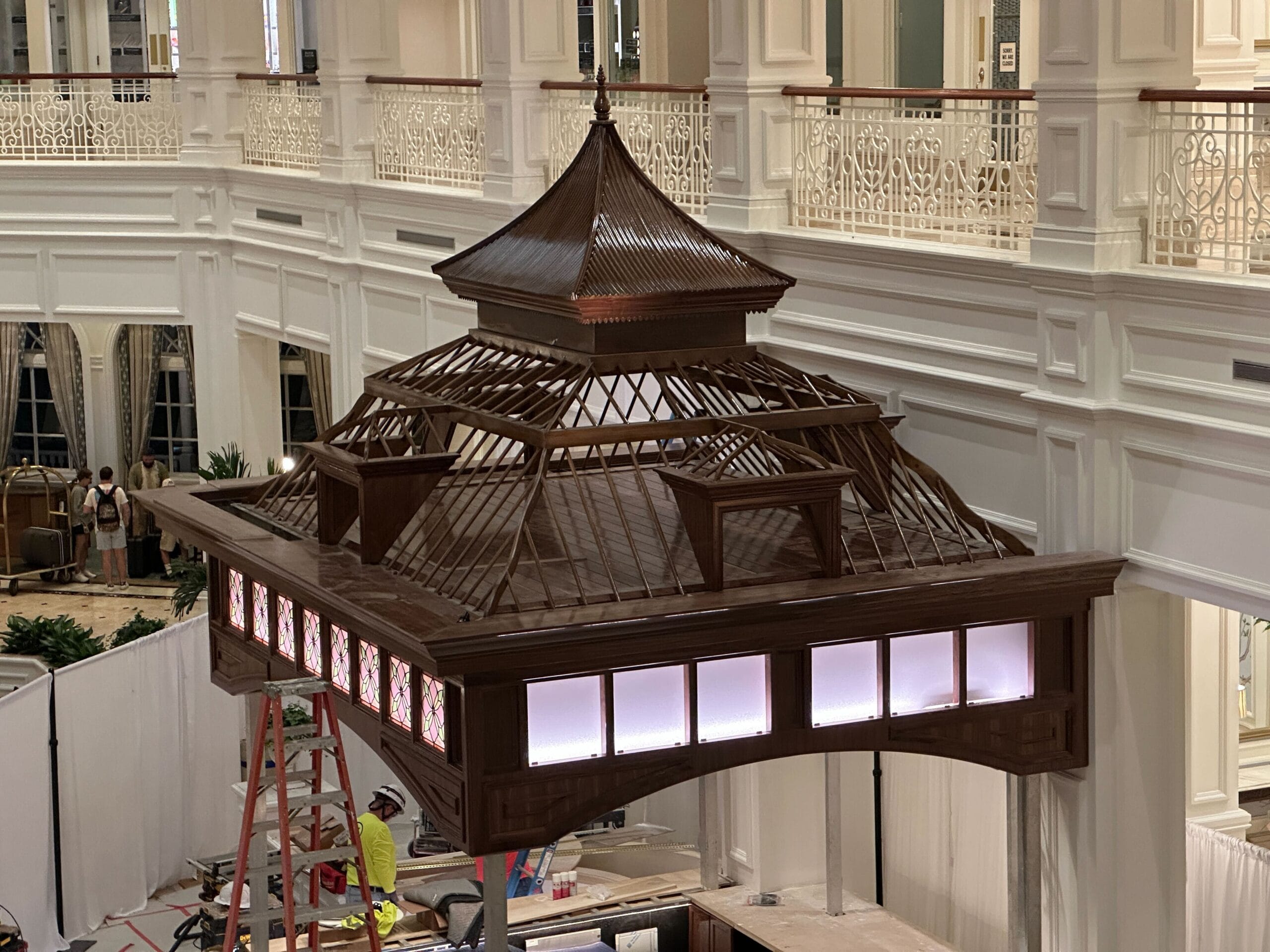 Large, ornate wooden pavilion suspended indoors, reminiscent of Disney park architecture; workers and white railings in background. Large, ornate wooden pavilion suspended indoors, reminiscent of Disney park architecture; workers and white railings in background.