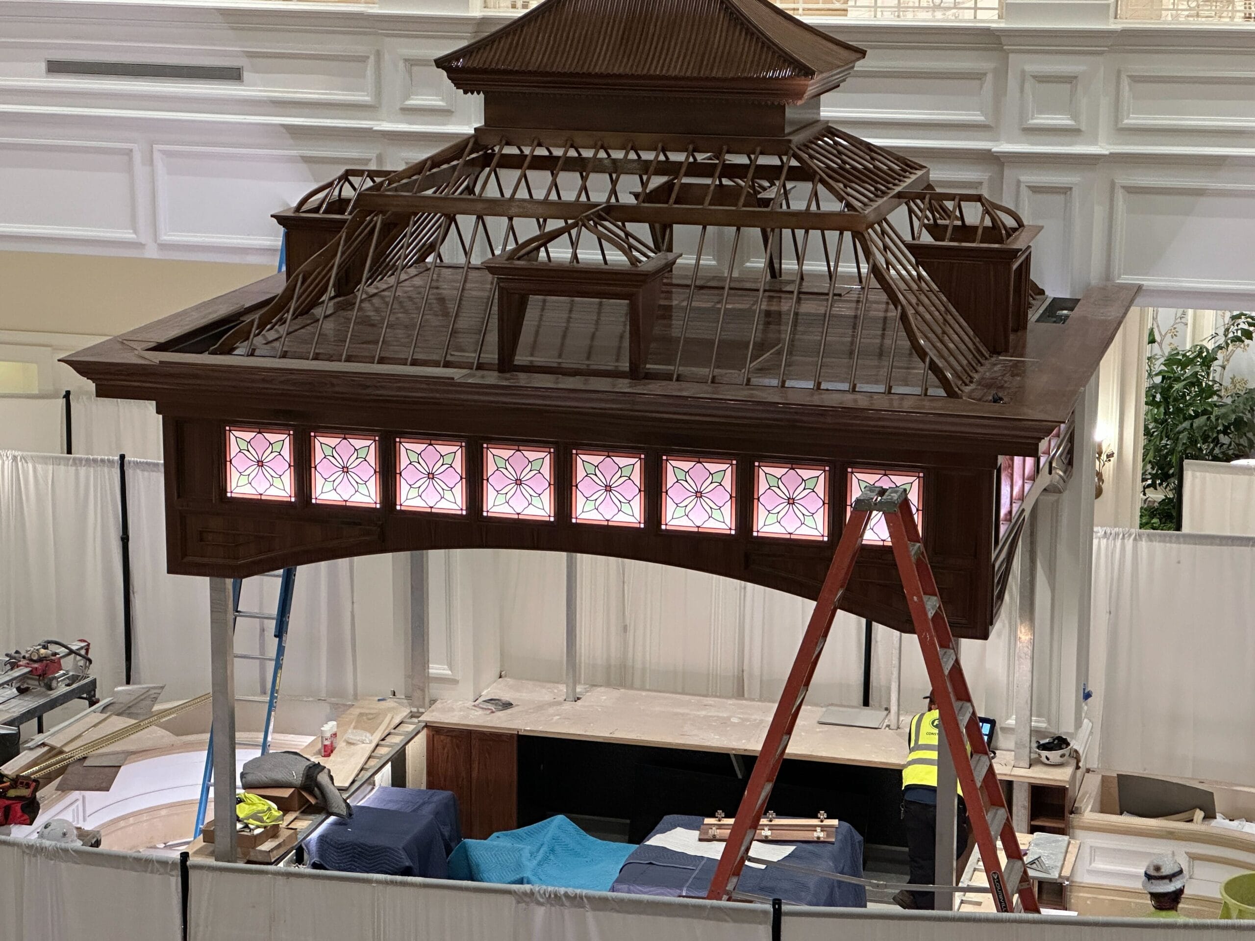 A worker on a ladder works next to a large wooden gazebo with stained glass panels, reminiscent of whimsical Disney park designs. A worker on a ladder works next to a large wooden gazebo with stained glass panels, reminiscent of whimsical Disney park designs.