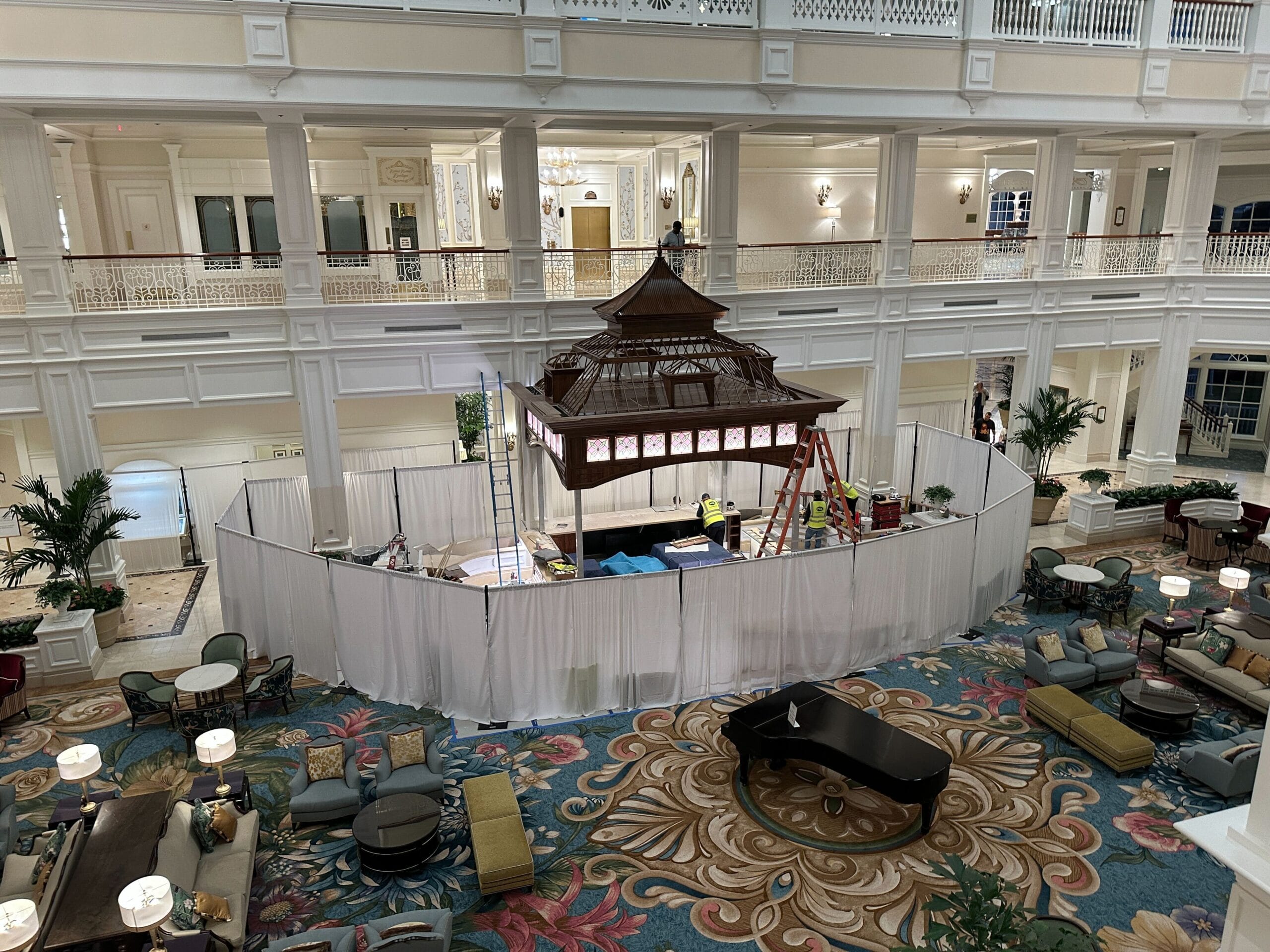 A busy Disney hotel lobby with a construction zone behind white curtains and workers around a gazebo, no castle visible. A busy Disney hotel lobby with a construction zone behind white curtains and workers around a gazebo, no castle visible.