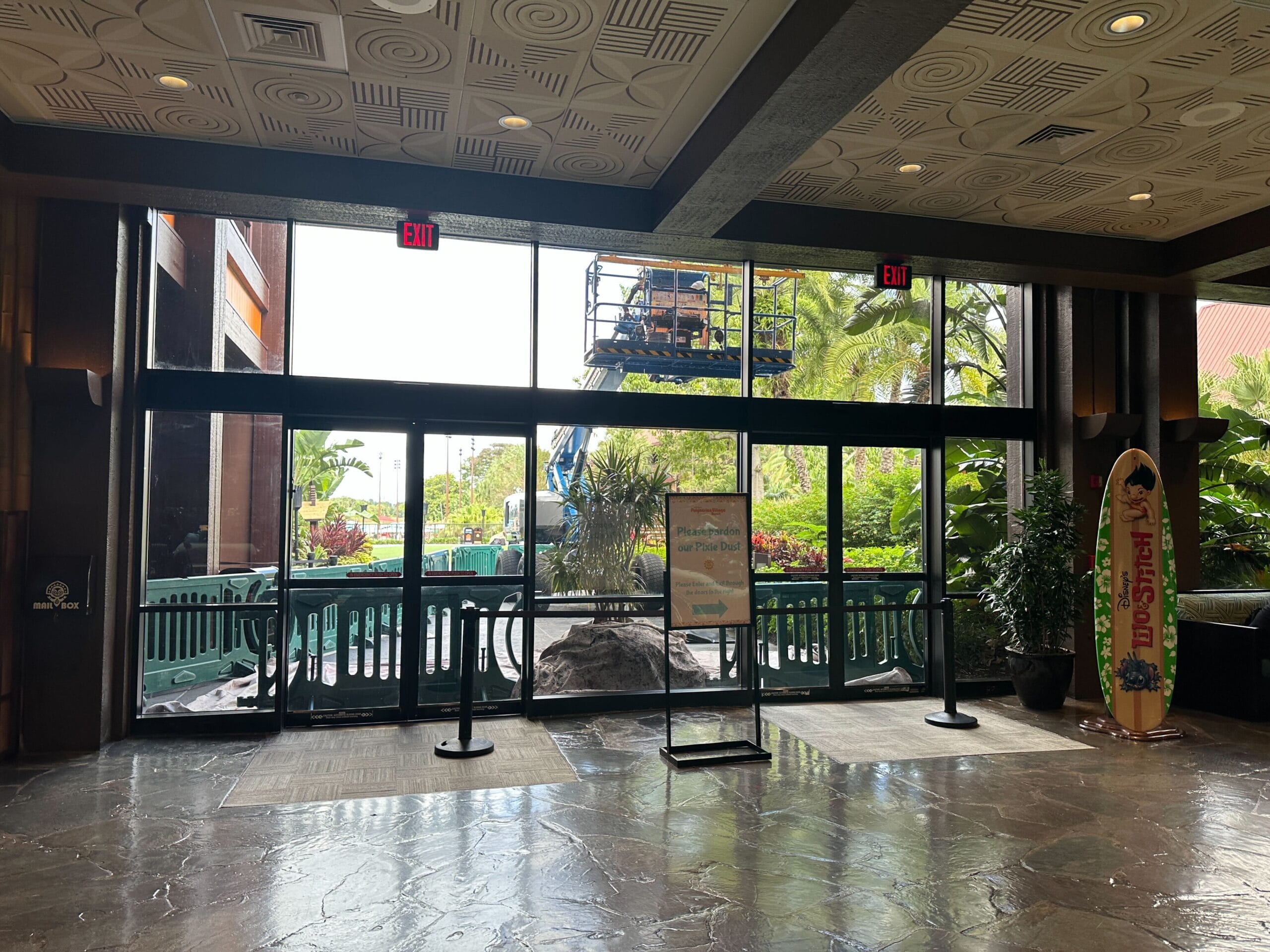 Disney resort lobby with large glass doors, tropical plants, a surfboard sign near the entrance, and a construction lift outside. Disney resort lobby with large glass doors, tropical plants, a surfboard sign near the entrance, and a construction lift outside.