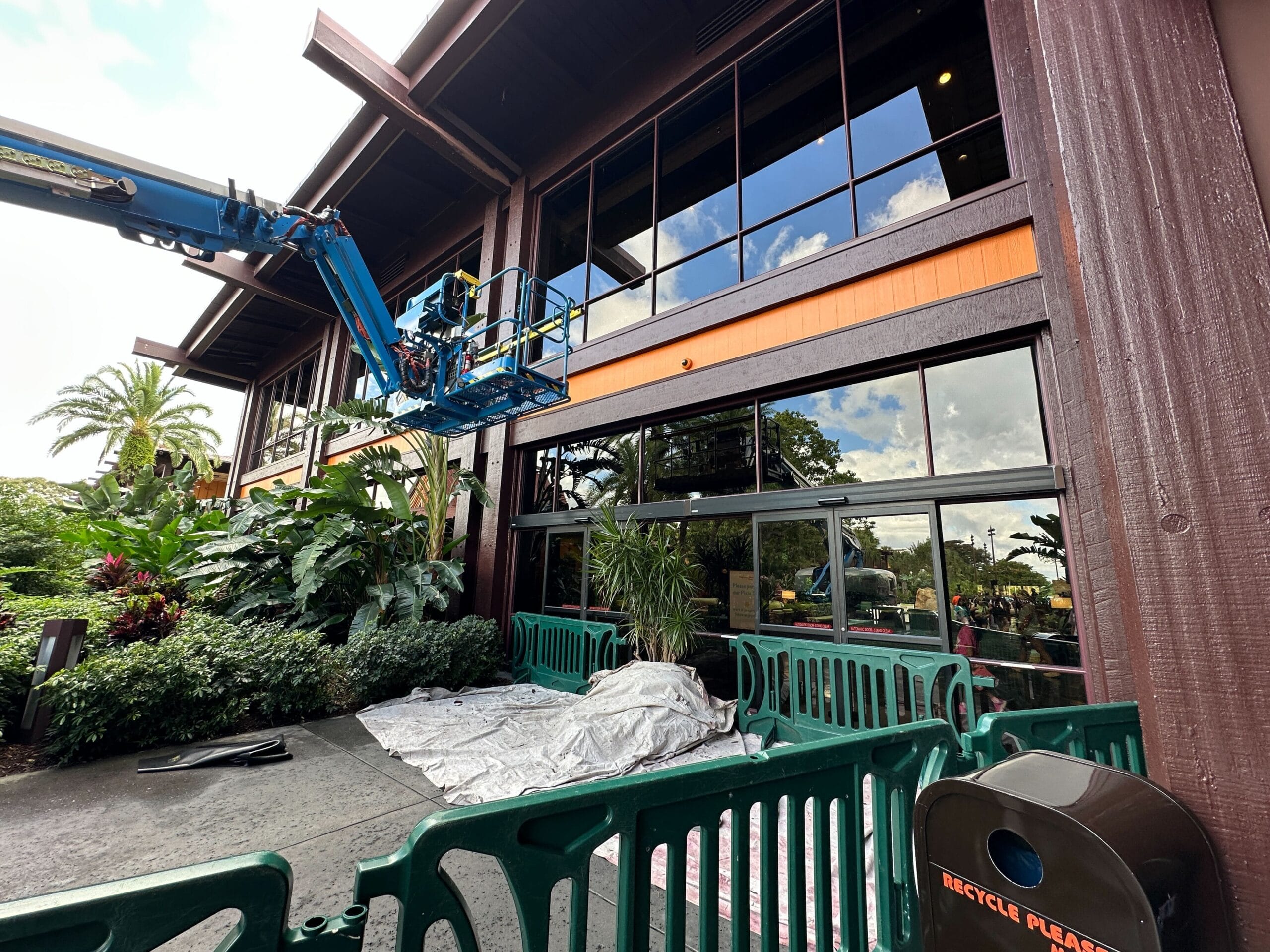 A worker repairs windows on a lift outside a building in a Disney park, with green barriers and themed landscaping around. A worker repairs windows on a lift outside a building in a Disney park, with green barriers and themed landscaping around.