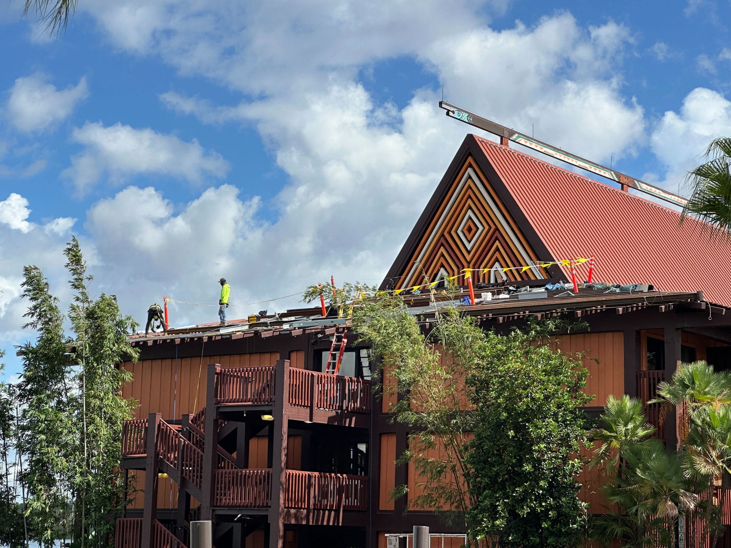 Workers in safety gear repair the roof of a brown building with a triangular facade, not resembling any Disney park castle, under a partly cloudy sky. Workers in safety gear repair the roof of a brown building with a triangular facade, not resembling any Disney park castle, under a partly cloudy sky.