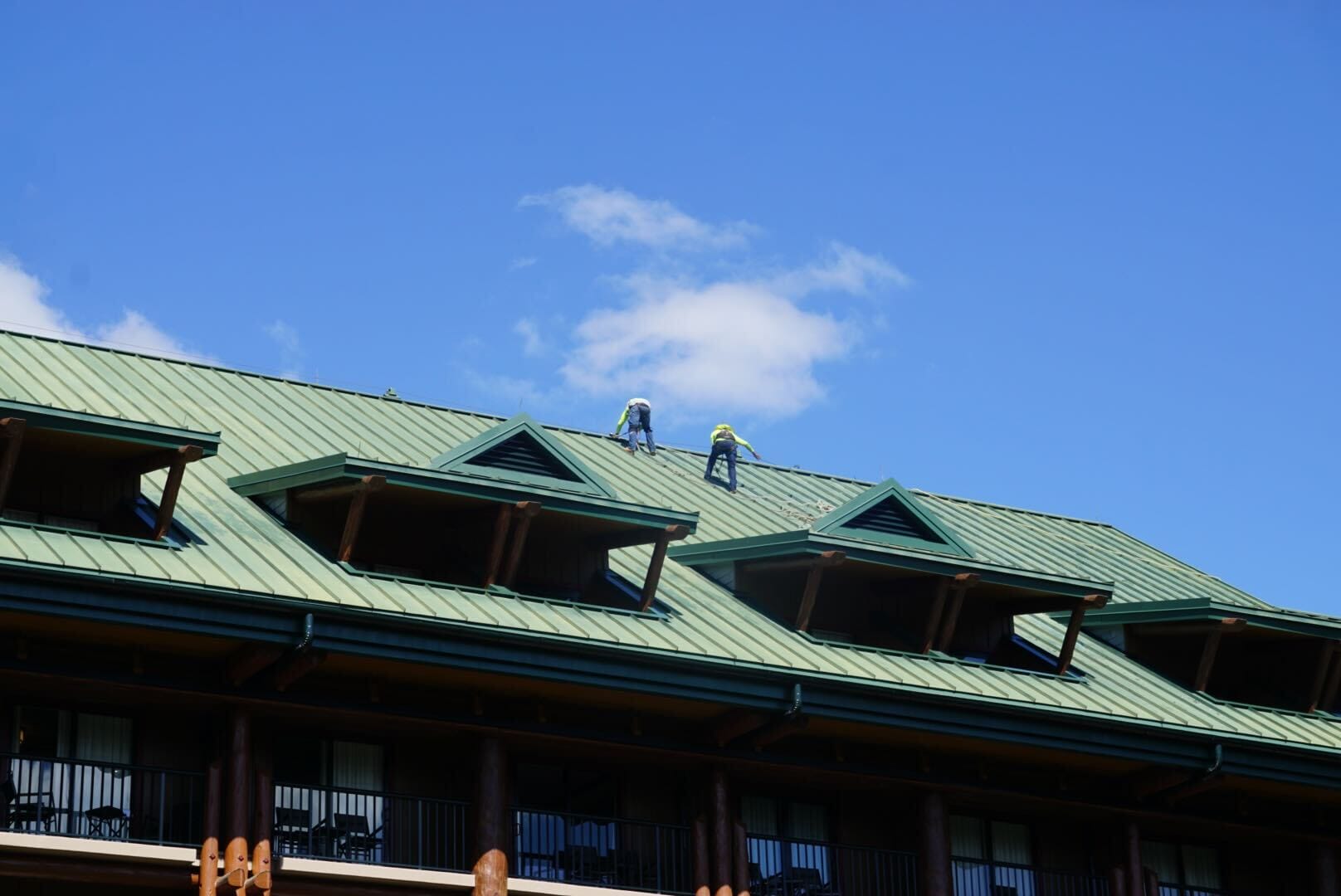 Two workers in safety gear work atop a green metal roof under a blue sky, reminiscent of rooftops seen in Disneyland parks. Two workers in safety gear work atop a green metal roof under a blue sky, reminiscent of rooftops seen in Disneyland parks.