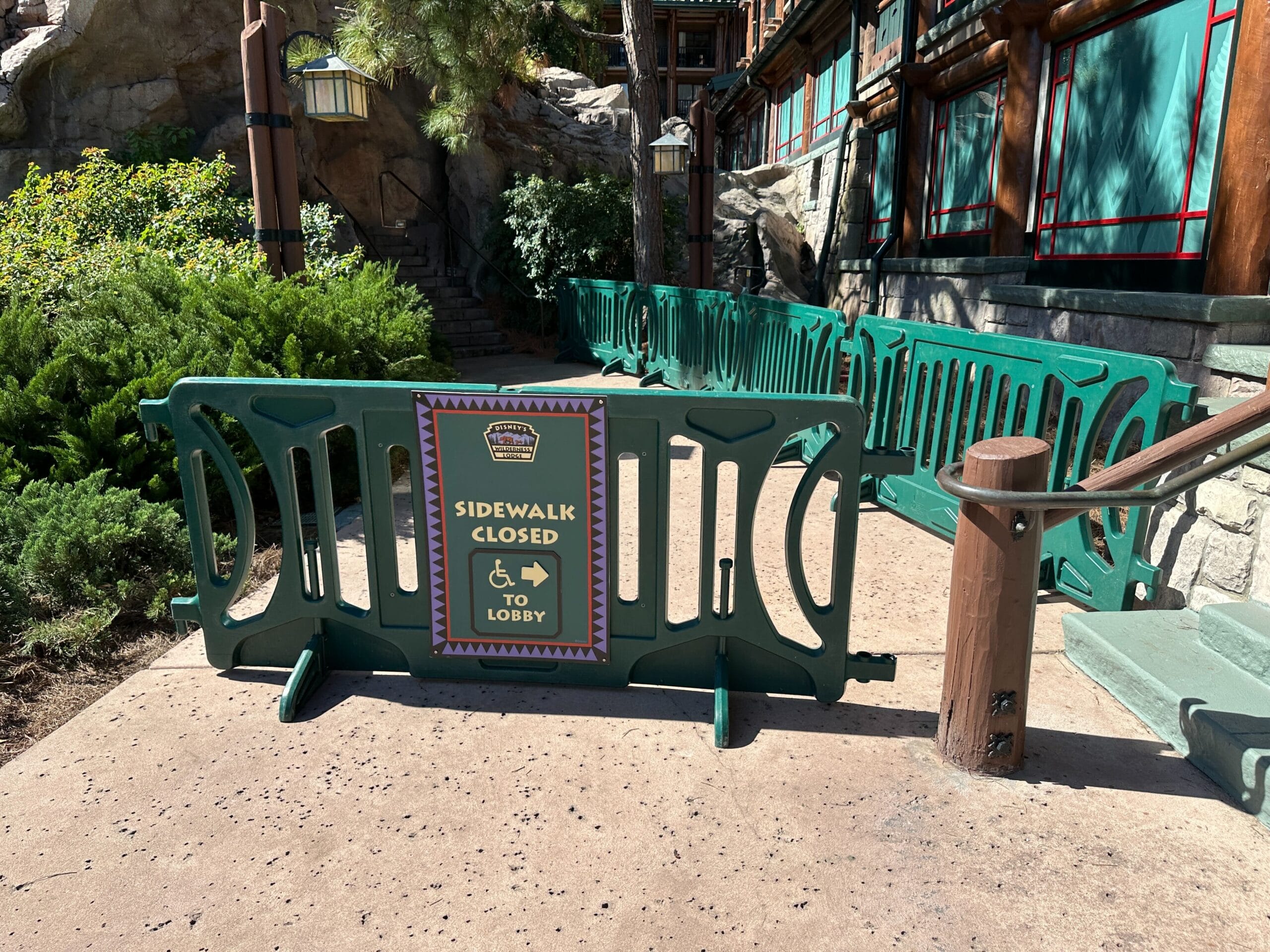 Green construction barriers block the sidewalk, with a sign saying Sidewalk Closed, to Lobby and a left arrow, Disney park style. Green construction barriers block the sidewalk, with a sign saying Sidewalk Closed, to Lobby and a left arrow, Disney park style.