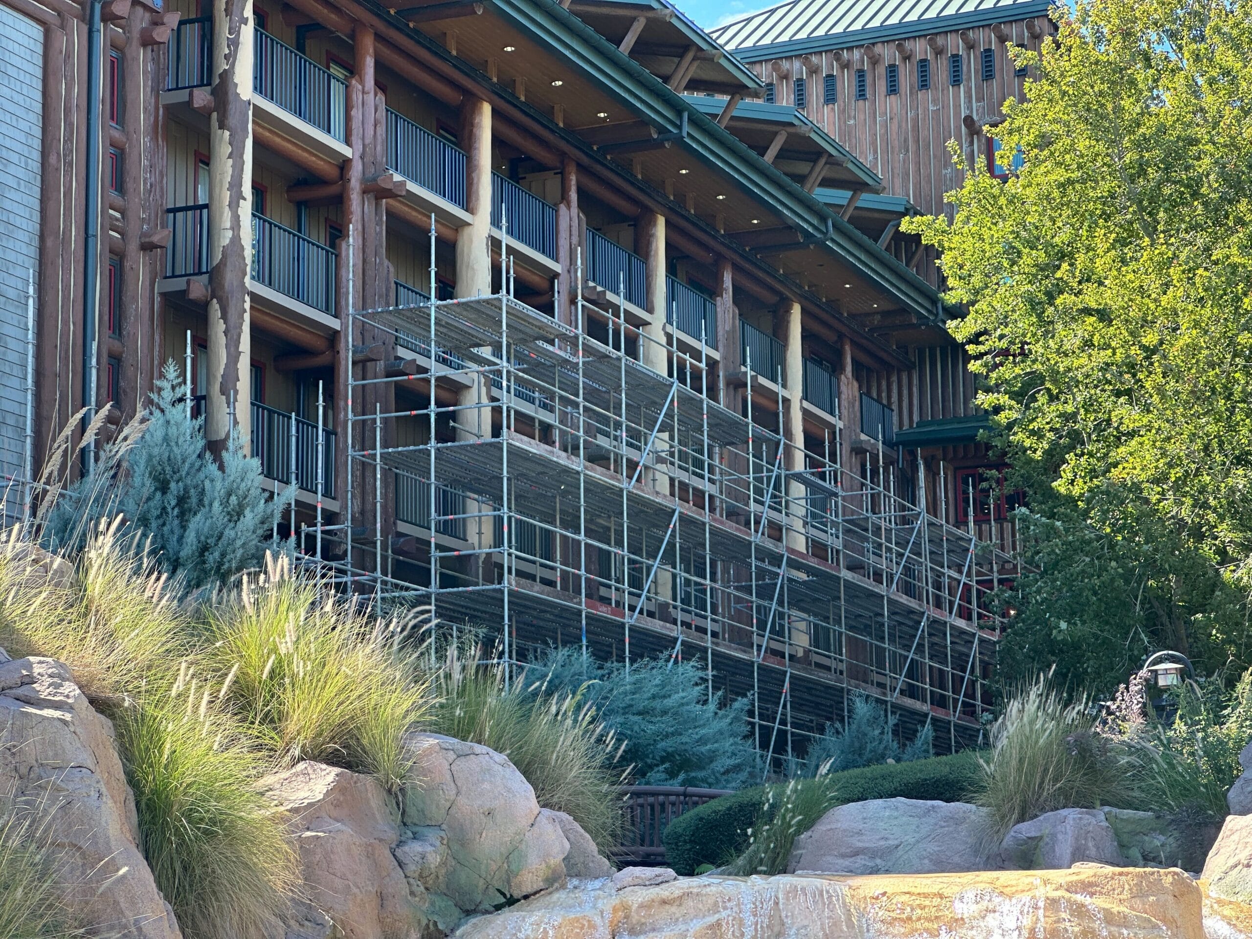 Scaffolding covers multiple floors of a rustic hotel, nestled among lush greenery and large rocks, reminiscent of Disney's Wilderness Lodge. Scaffolding covers multiple floors of a rustic hotel, nestled among lush greenery and large rocks, reminiscent of Disney's Wilderness Lodge.